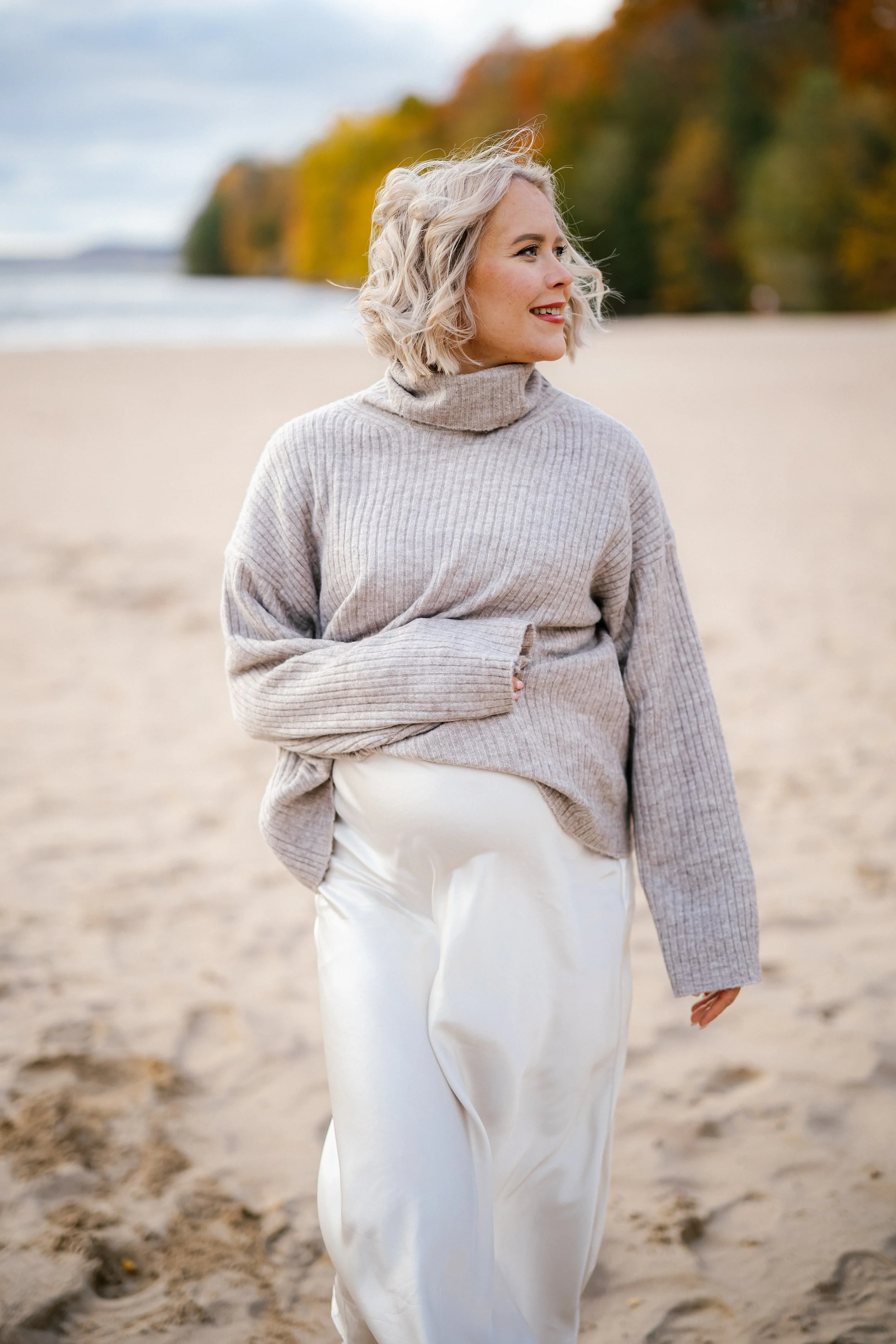 A woman with blonde, wavy hair standing on a sandy beach, wearing a beige ribbed turtleneck sweater and white satin pants, smiling and looking to the side with a background of trees with fall foliage.