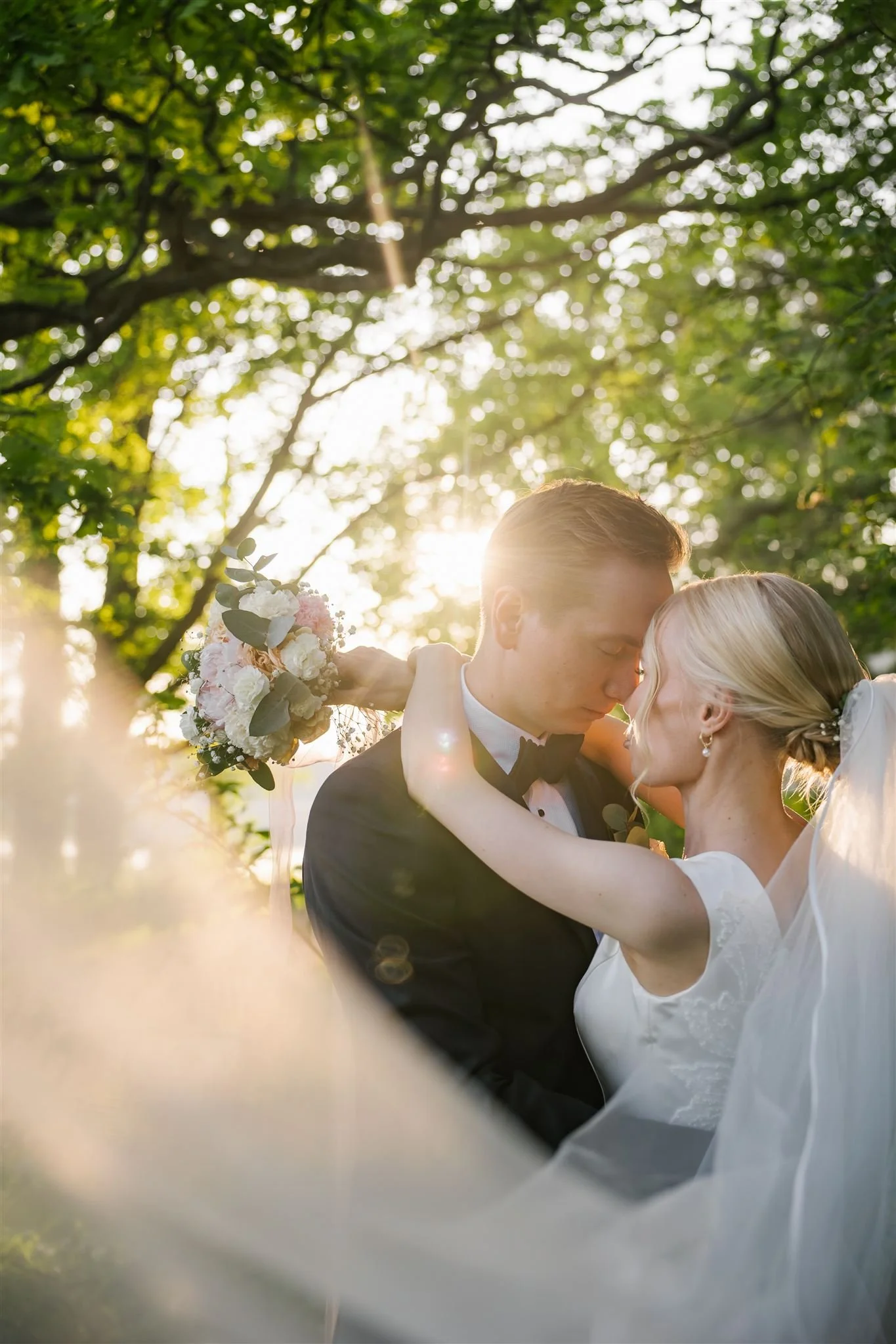 A bride and groom embracing outdoors during sunset with trees and sunlight in the background.