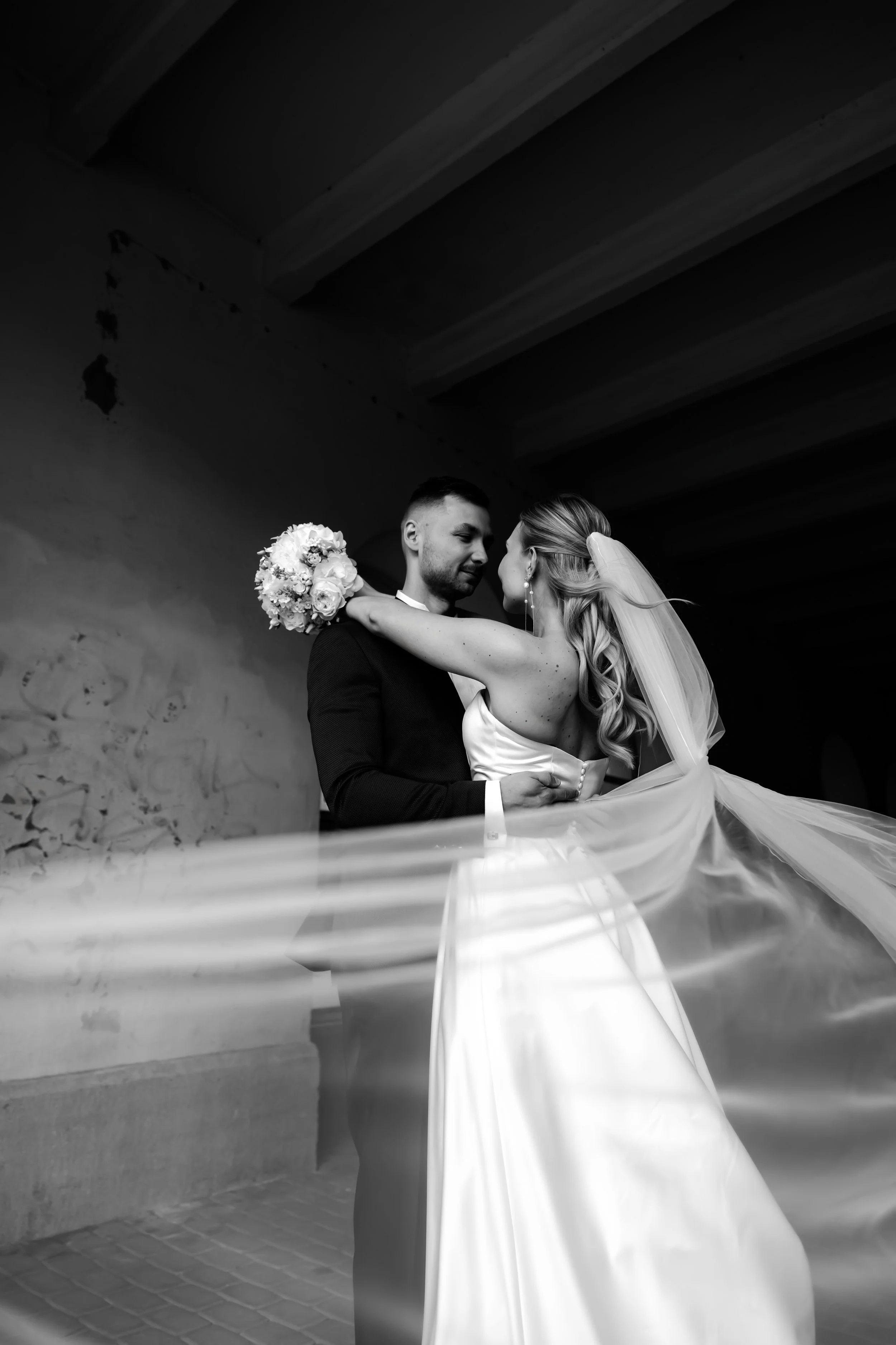 A black and white photo of a bride and groom sharing a dance, with the bride holding a bouquet of flowers and her veil flowing over the groom. They are close together, smiling, under a dark ceiling.