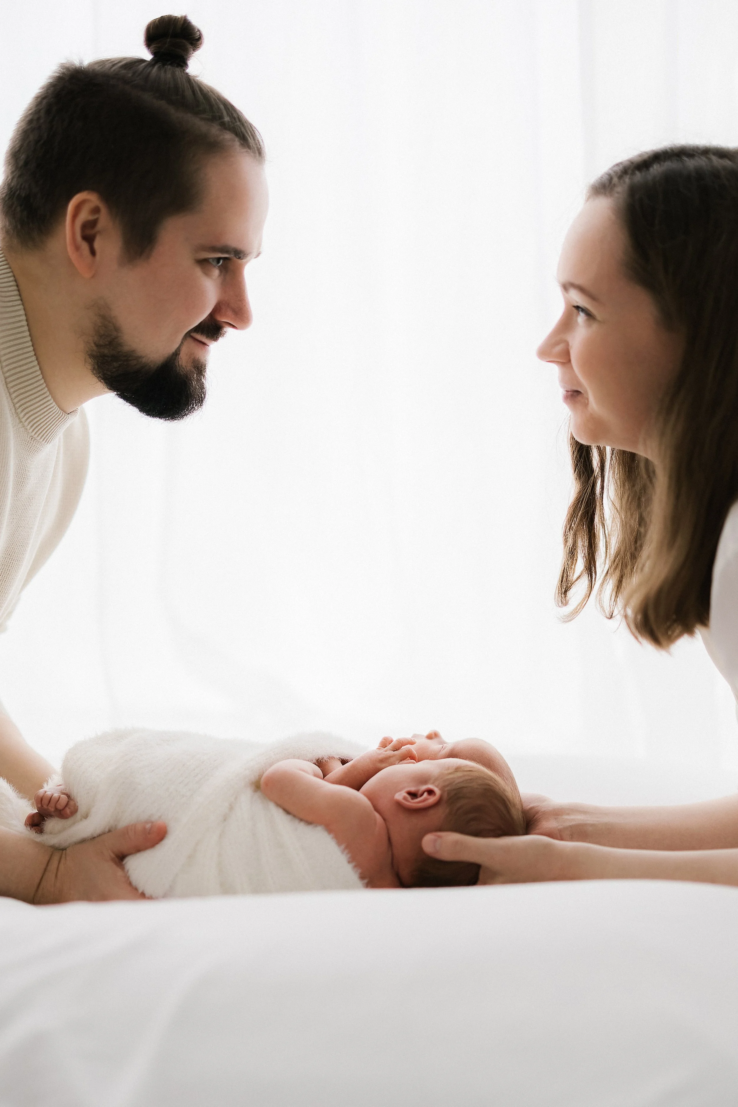 A man and woman are leaning towards each other, holding a newborn baby wrapped in a white blanket, with soft natural light in the background.