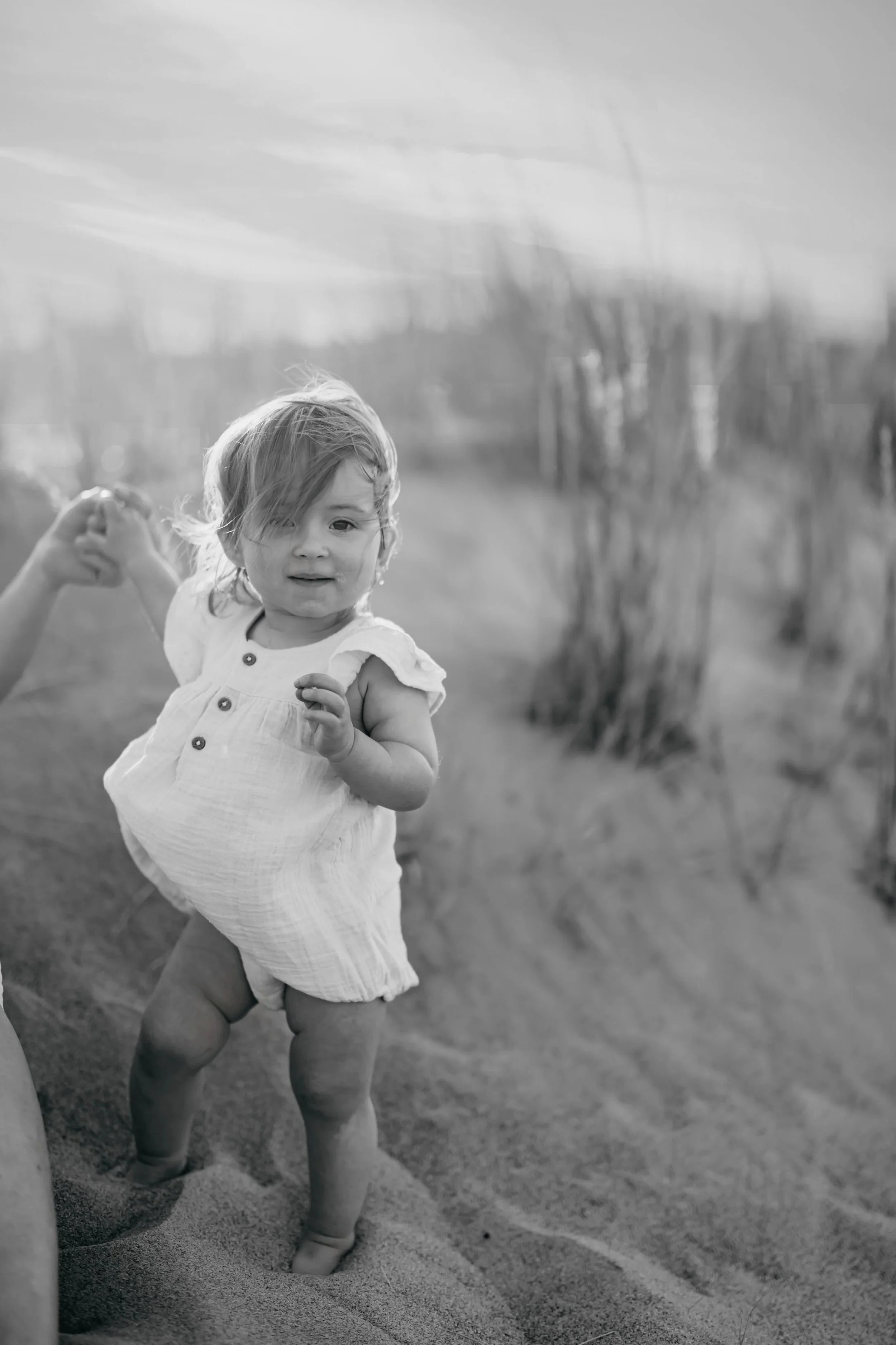 A young child standing on sandy ground, holding an adult's hand, with a blurred natural background, in black and white.