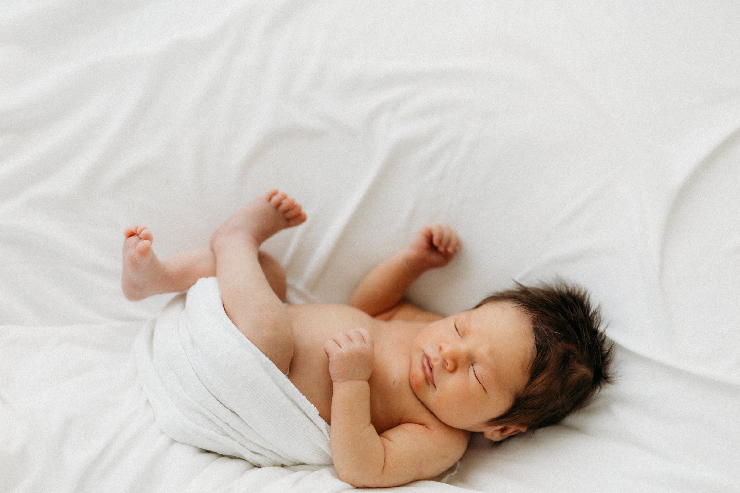 A newborn baby sleeping on a white bed, wrapped loosely in a white cloth.