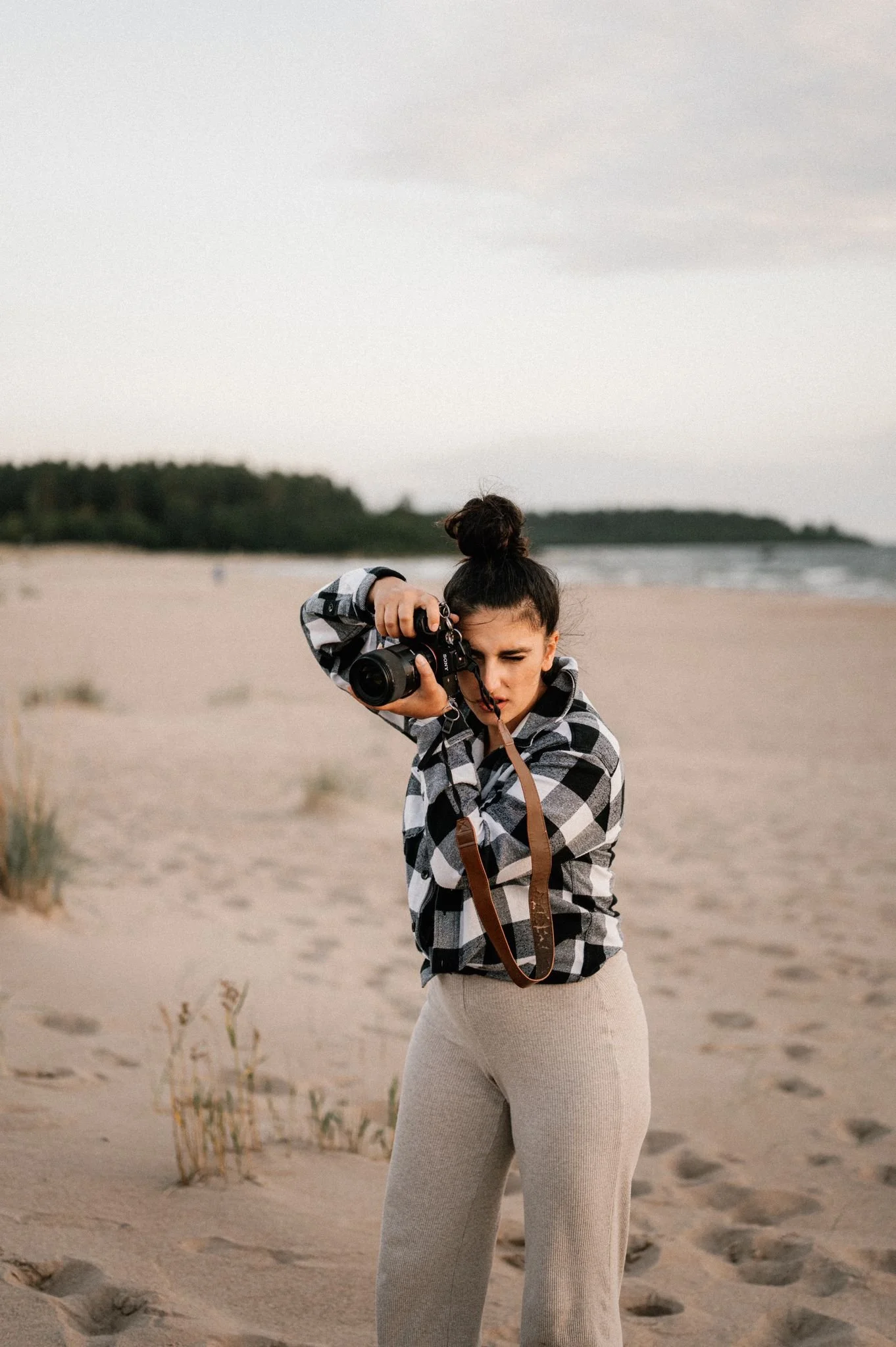 Young woman with dark hair in a bun, wearing a black and white checkered jacket and light-colored pants, taking a photo with a camera on a sandy beach during sunset or overcast weather.