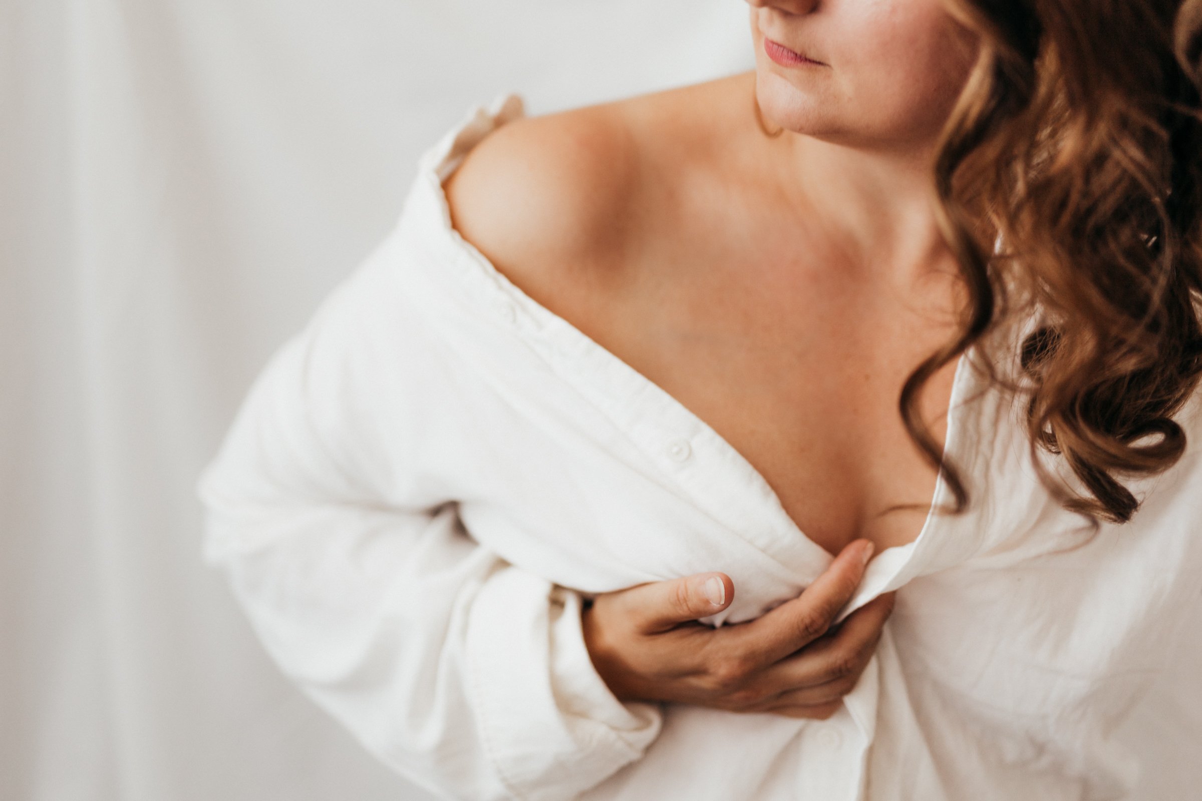 Woman wearing a white shirt holding her chest, partially unbuttoned, revealing her shoulder and collarbone.