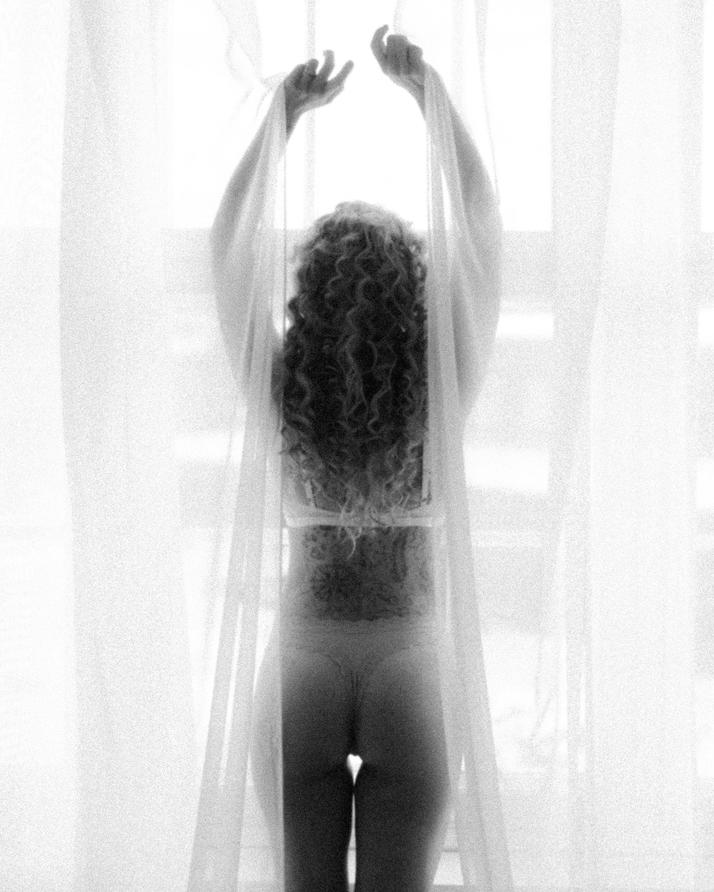 A woman with long curly hair standing behind sheer curtains, viewed from behind, wearing minimal clothing.