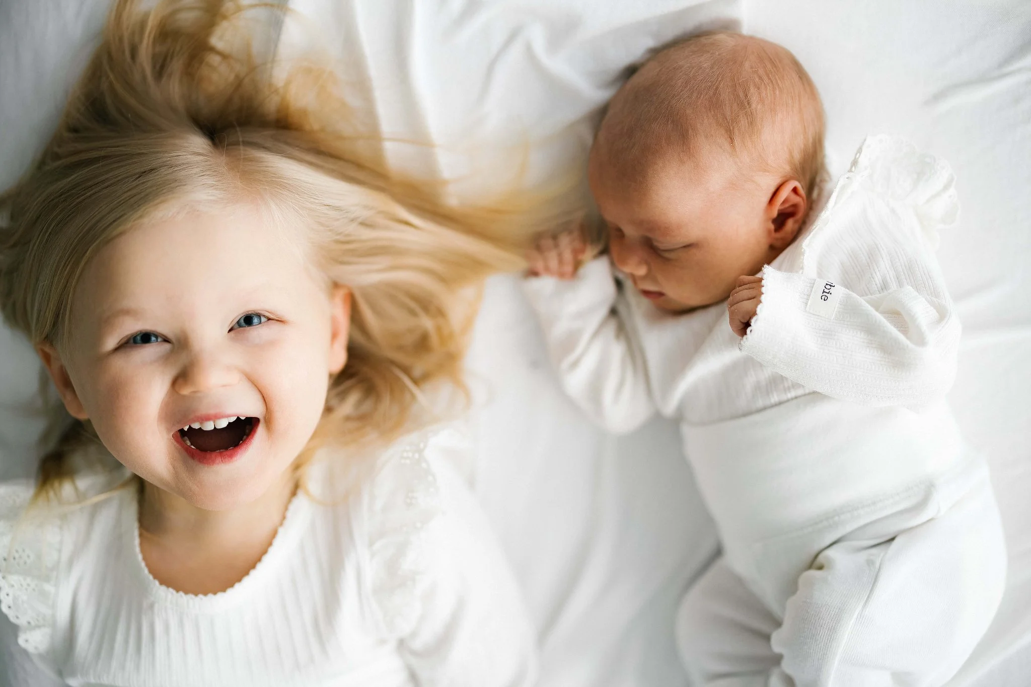 Two young children, a girl and a baby, lying on a bed with white sheets, smiling and looking at the camera.