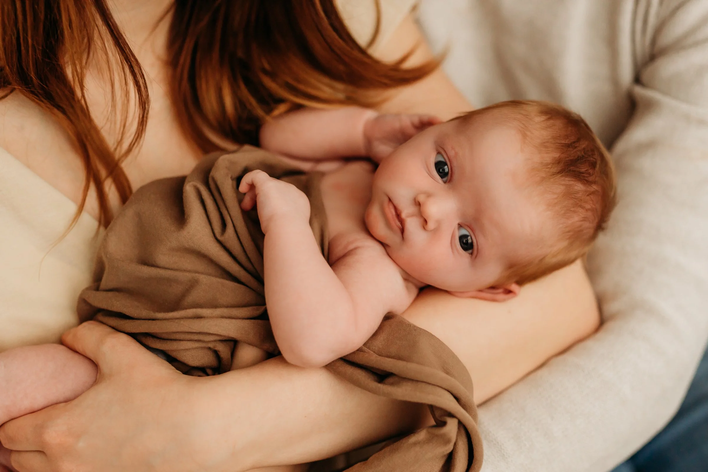 Close-up of a woman with red hair holding a sleeping baby with fair skin and brown hair, wrapped in a brown blanket, on a light-colored pillow.