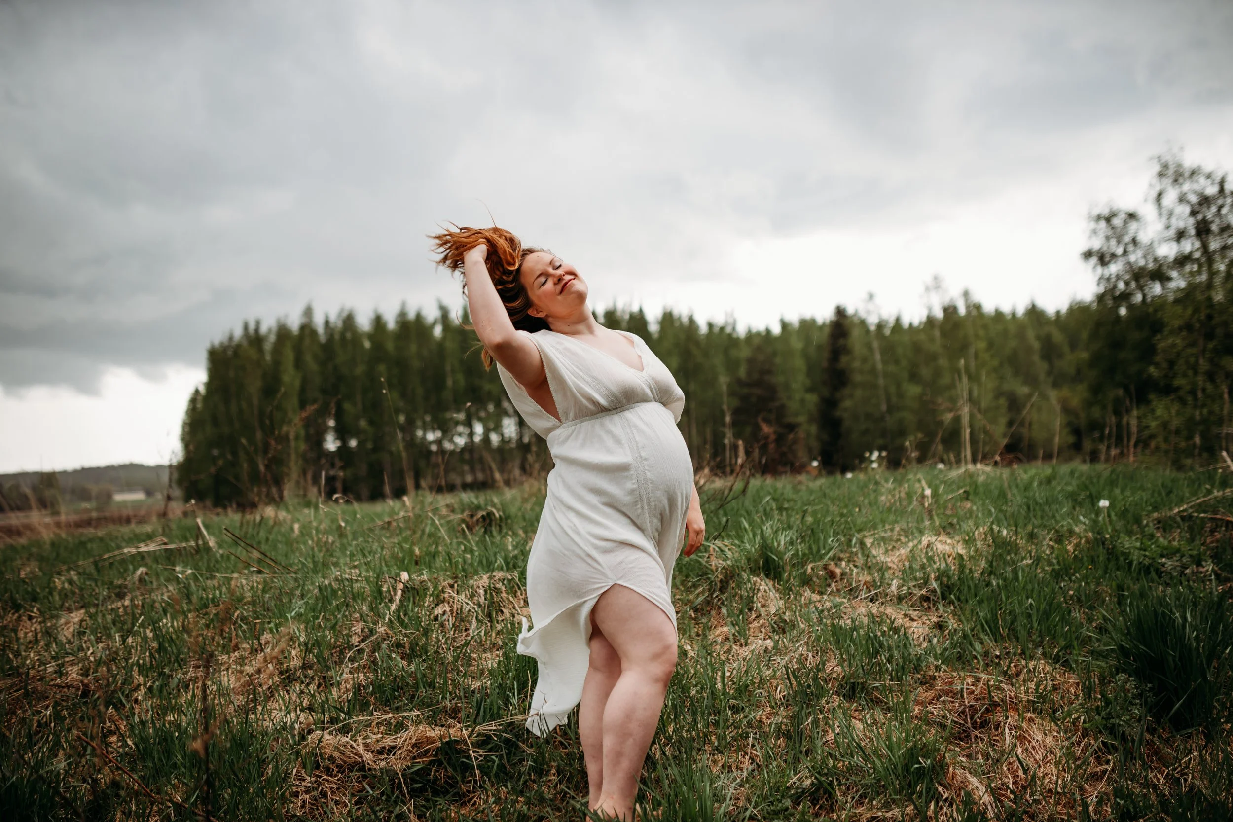 A pregnant woman in a white dress standing in a grassy field with a forest in the background, tossing her hair back and smiling under a cloudy sky.