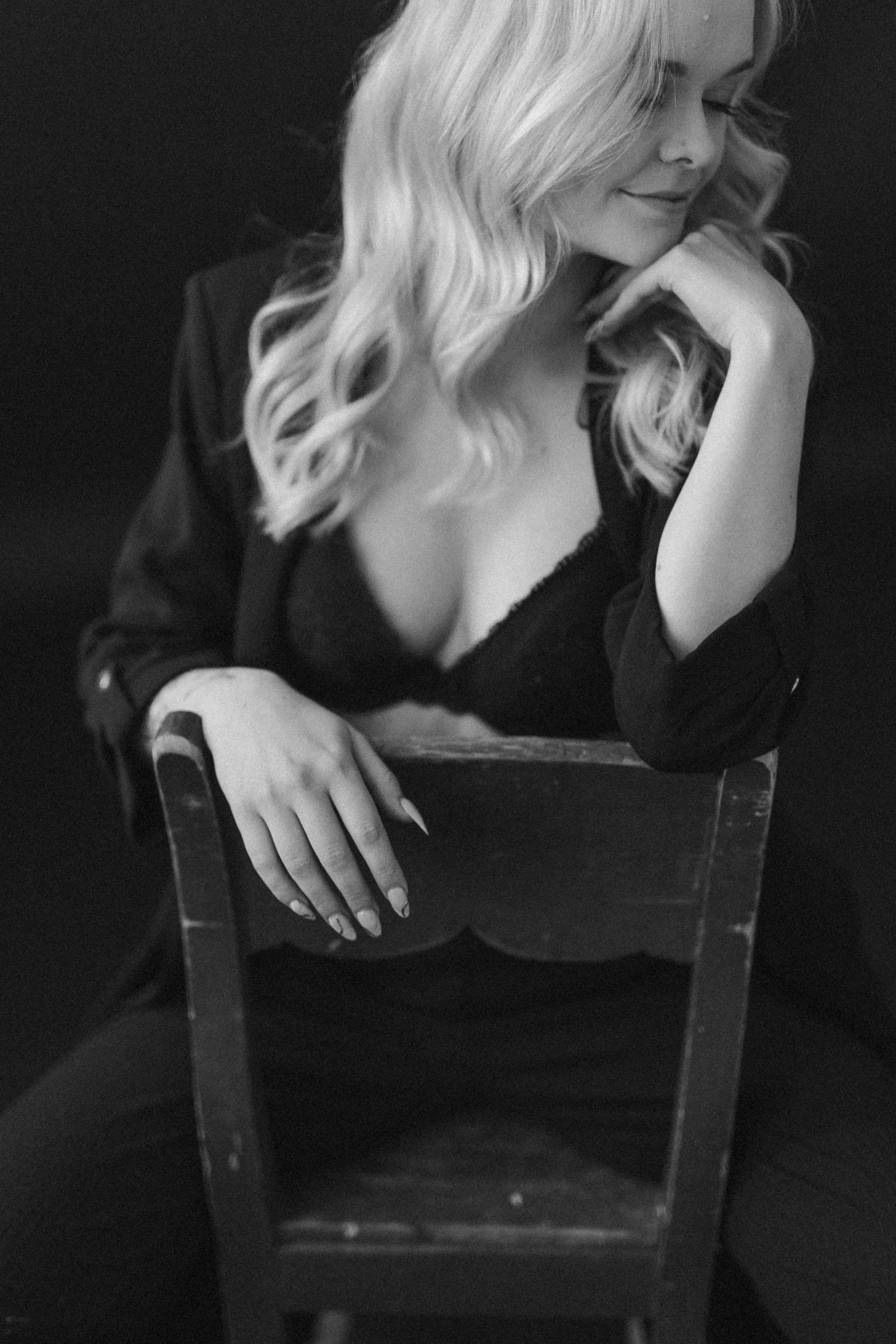 A black and white photo of a woman with wavy blonde hair, wearing a black lace top and a jacket, sitting on a chair with her hand resting on the chair's back, her eyes closed and smiling softly.