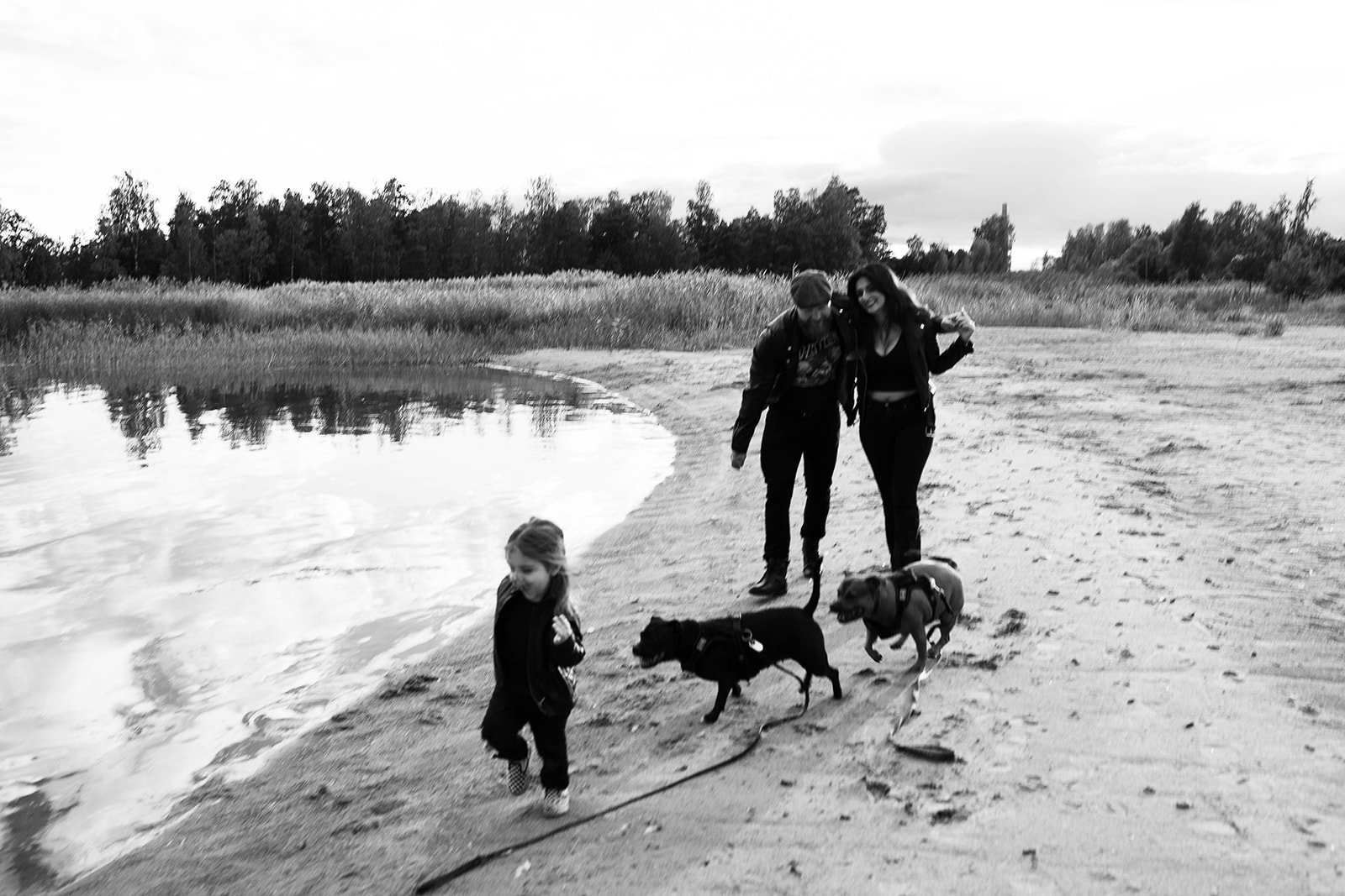 A family of four with two dogs walking along a beach next to a body of water, with trees and grass in the background, in black and white.