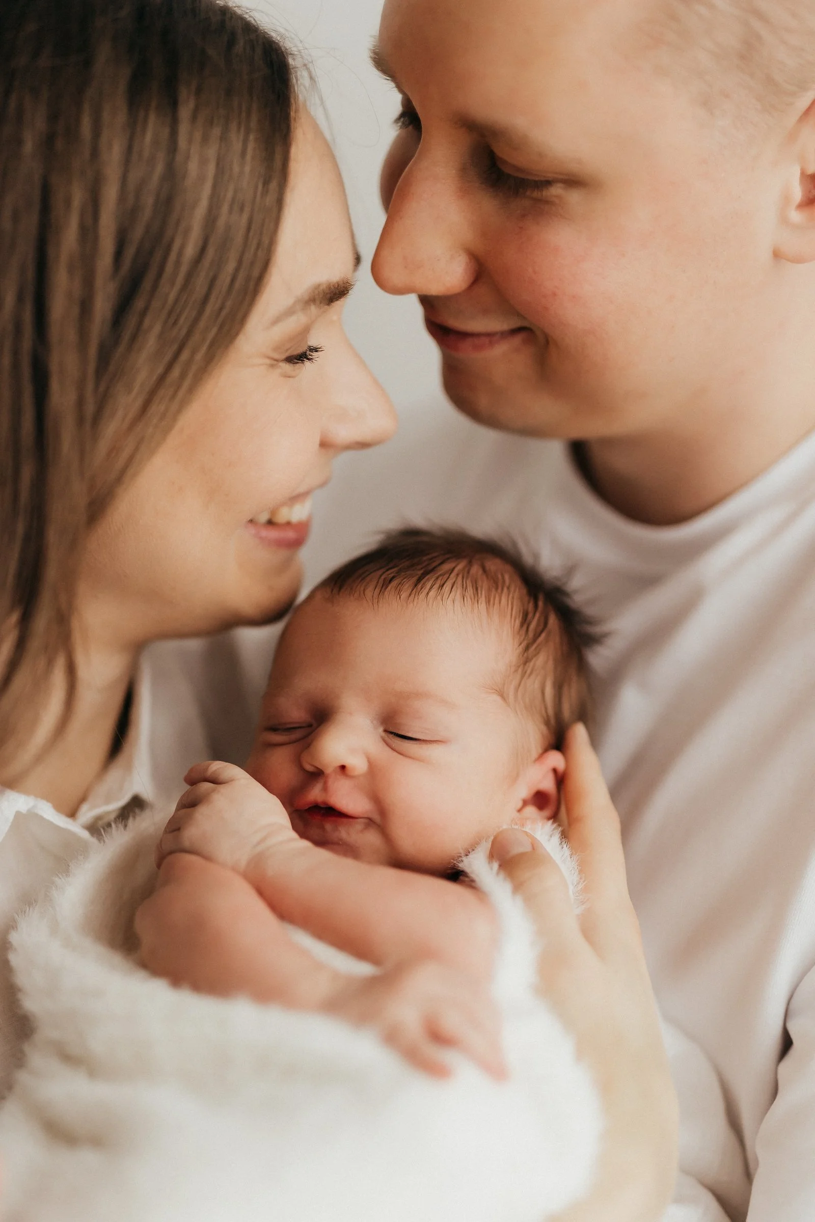 A newborn baby being held and cradled by a woman and a man, all smiling and close together, with the baby's eyes closed.