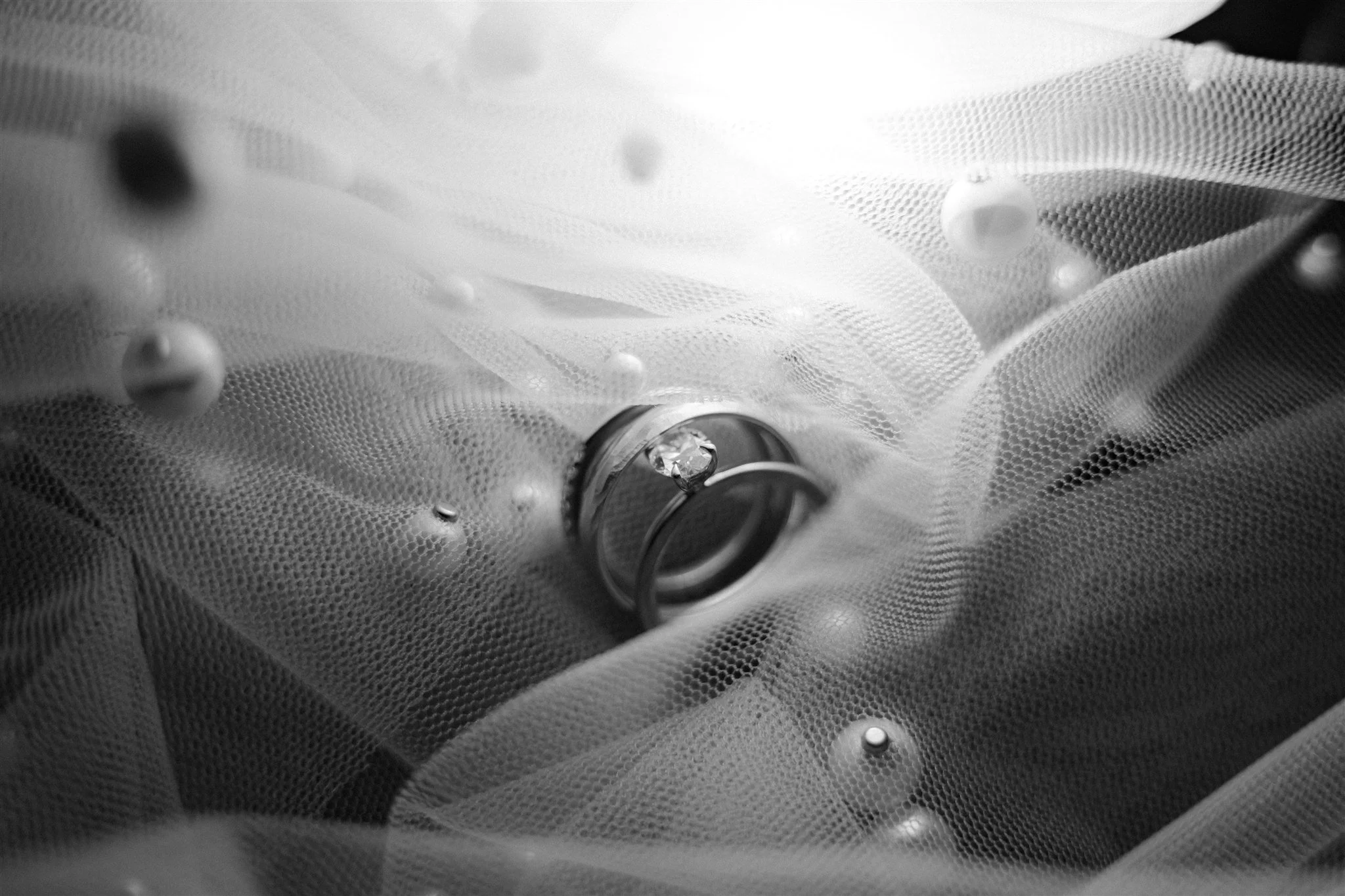 A close-up black and white photograph of a diamond ring with a wedding band, placed on a textured fabric with decorative beads scattered around.