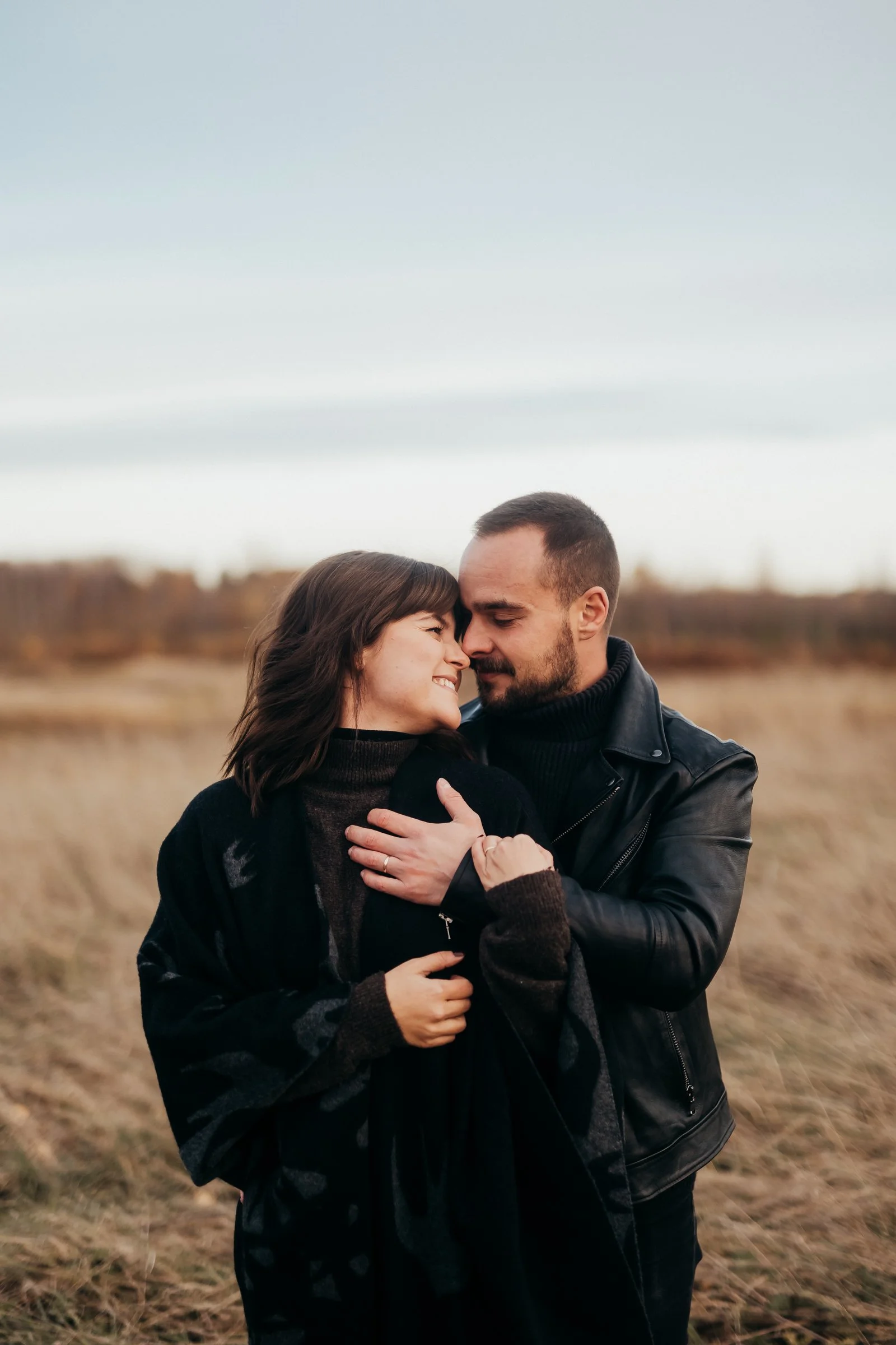 A woman and a man are embracing outdoors, touching foreheads, smiling and showing affection. The woman has shoulder-length brown hair and the man has short dark hair and a beard, wearing a black leather jacket.