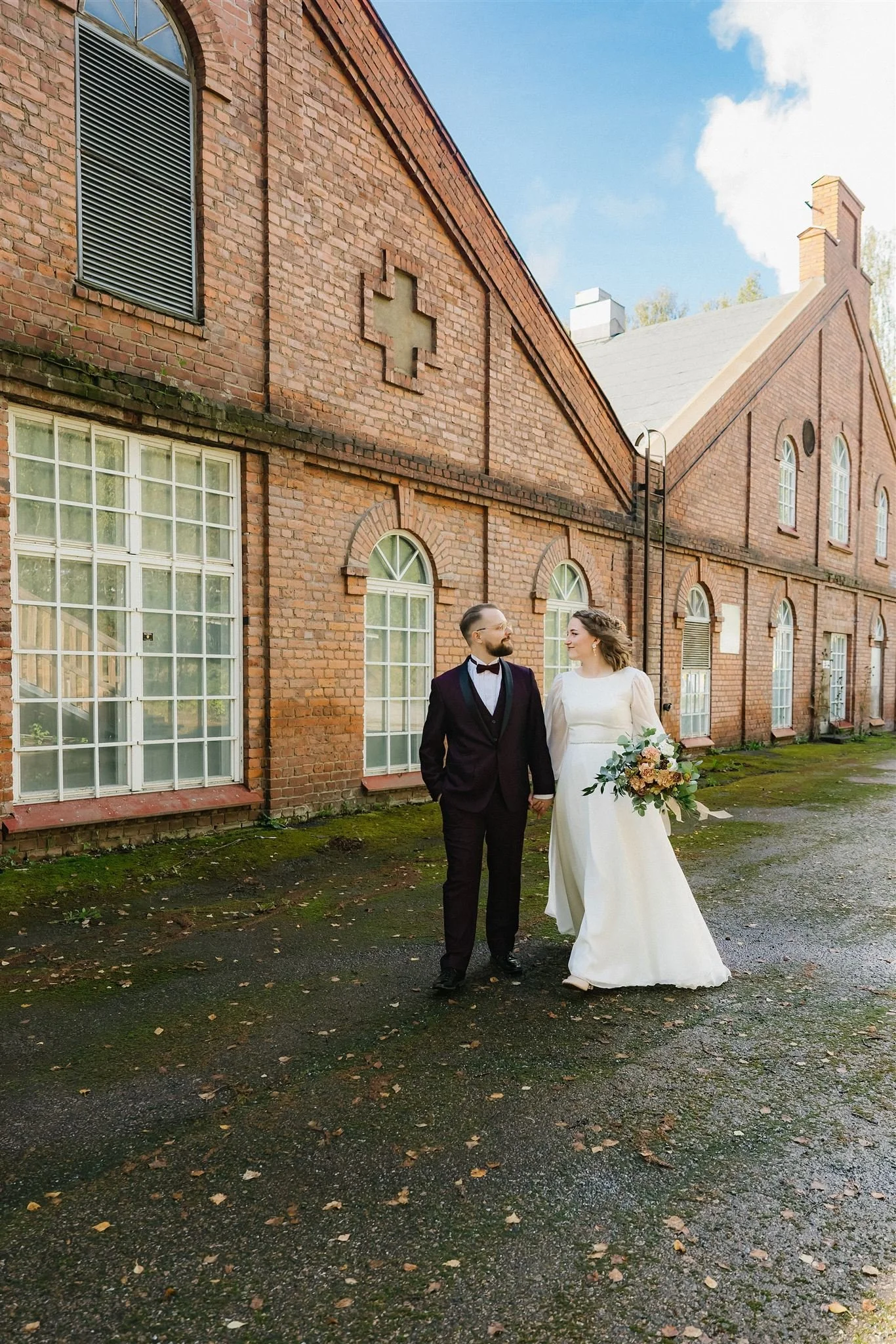 A bride and groom walking hand in hand outside of a brick building with large arched windows and a cross design on the wall, celebrating their wedding day.