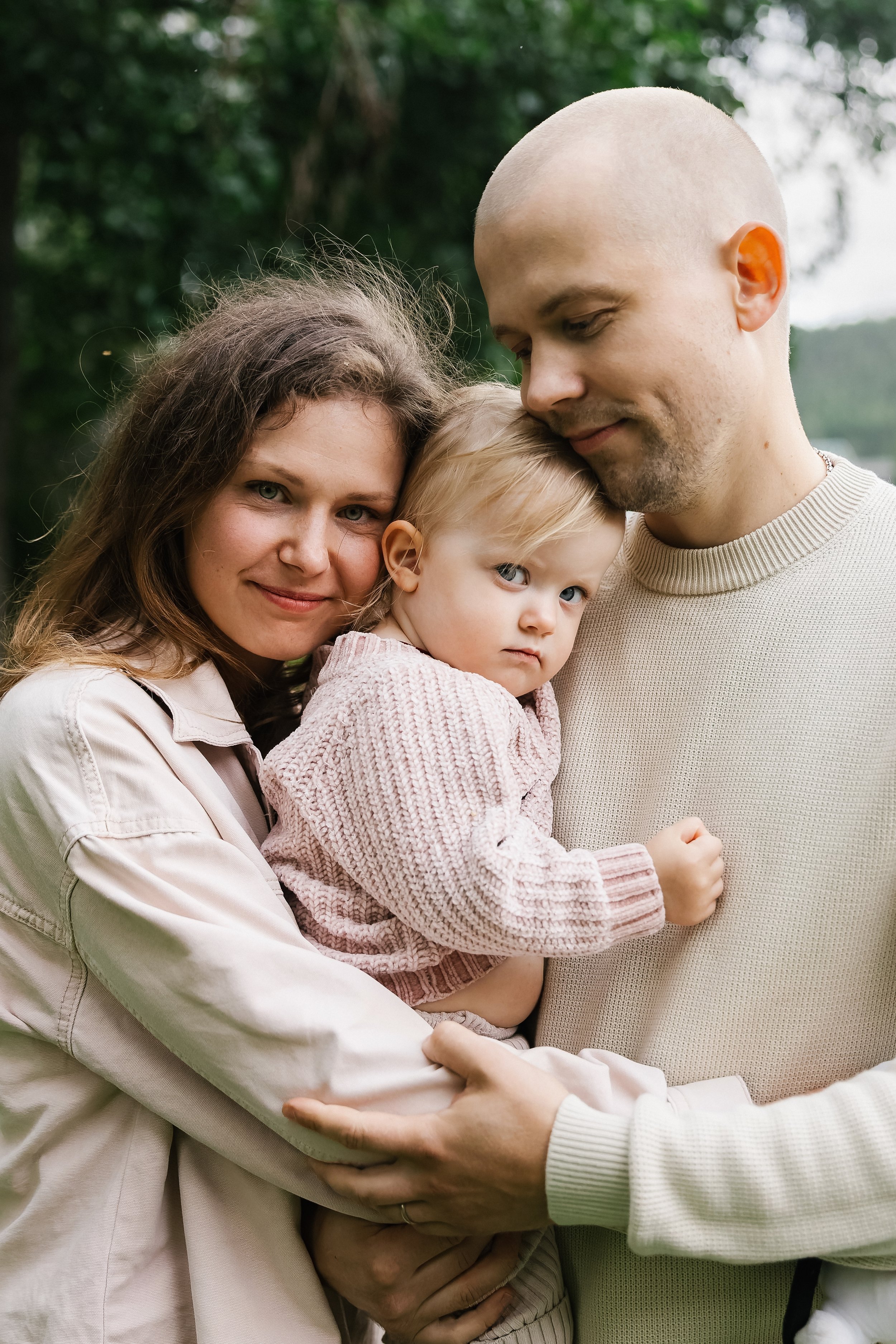 A family of three outdoors, a woman, a man, and a young girl, hugging each other, with trees and greenery in the background.