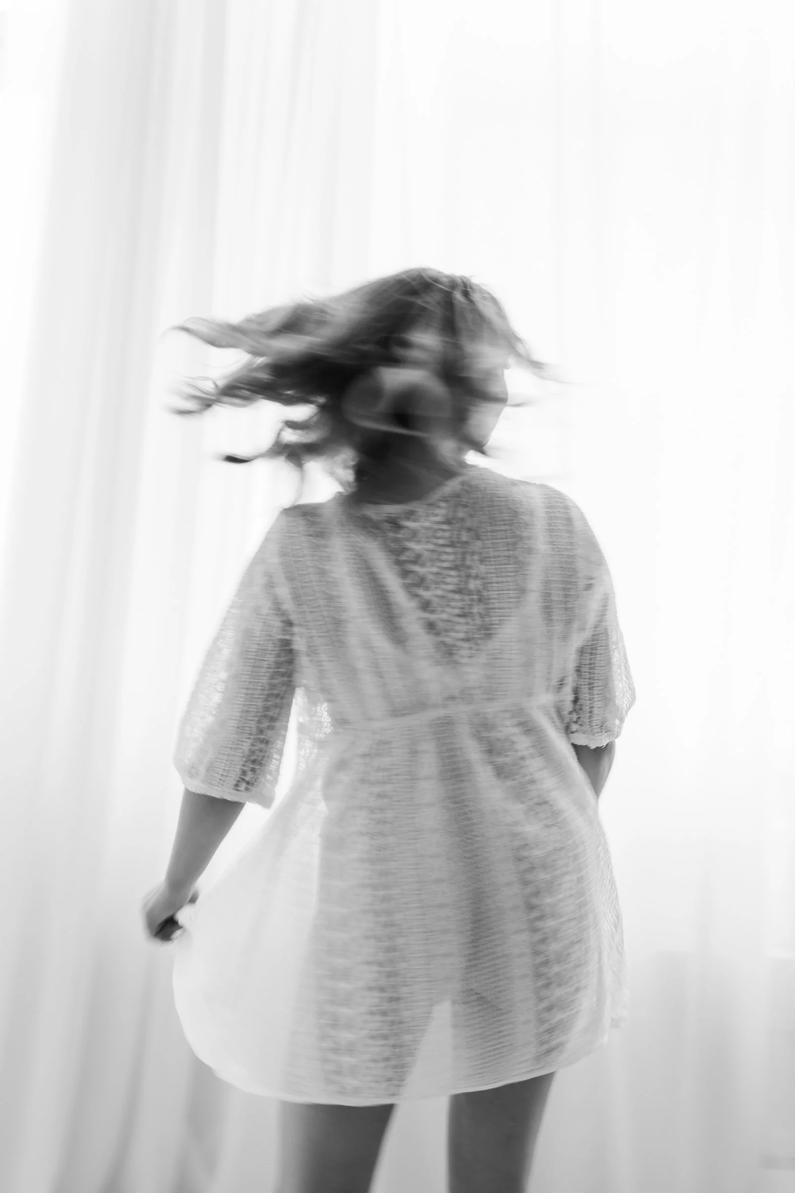 Black and white photo of a woman with long, wavy hair turning to the side, wearing a loose dress, standing in front of a light-colored curtain.