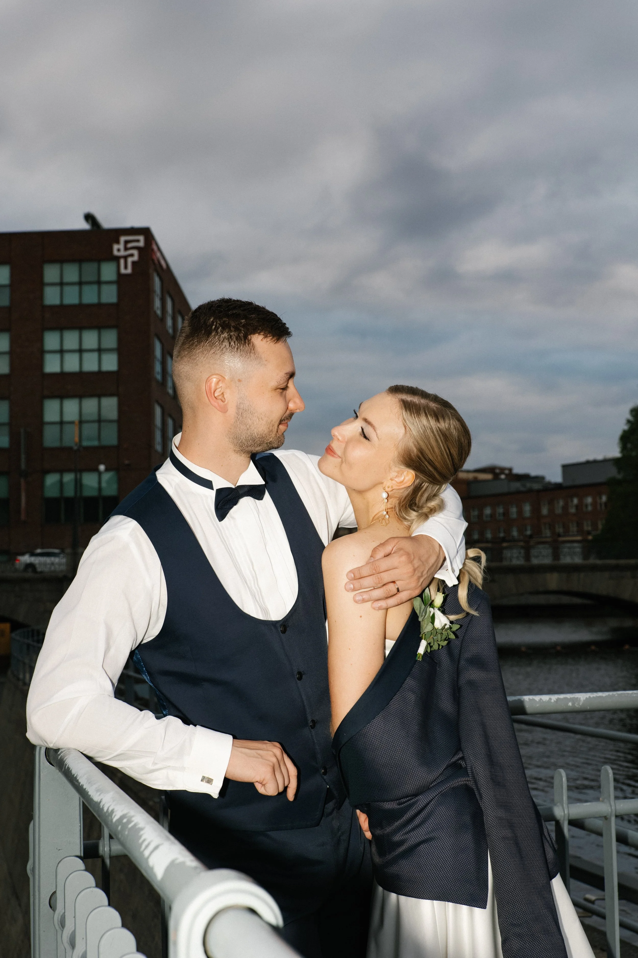 A newly married couple in formal attire sharing a romantic moment outdoors near a river during cloudy weather.