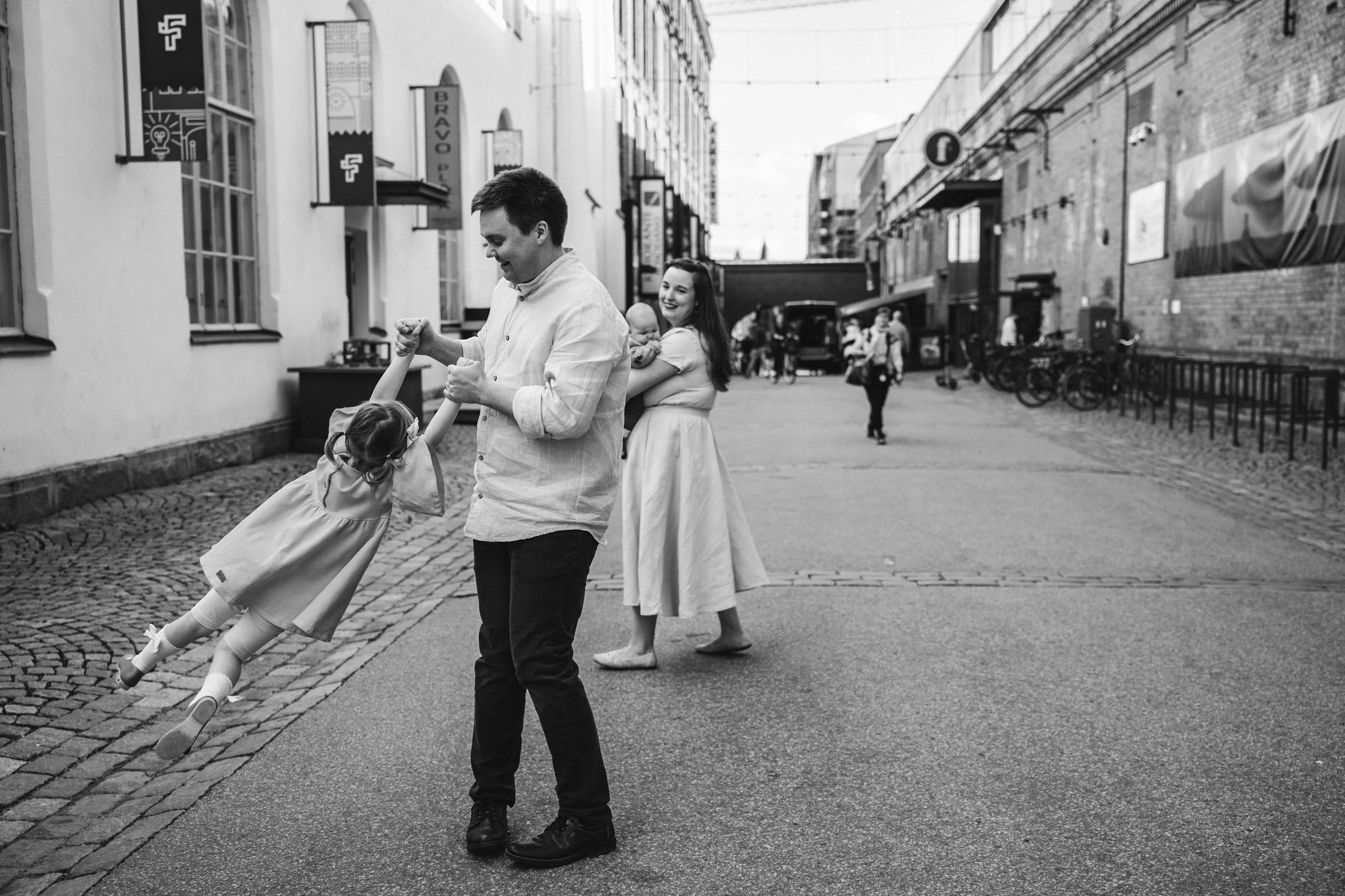 A black and white photo of a man spinning a young girl by her hands on a cobblestone street. A woman holding a baby watches and smiles in the background. Buildings and bicycles are visible along the street.