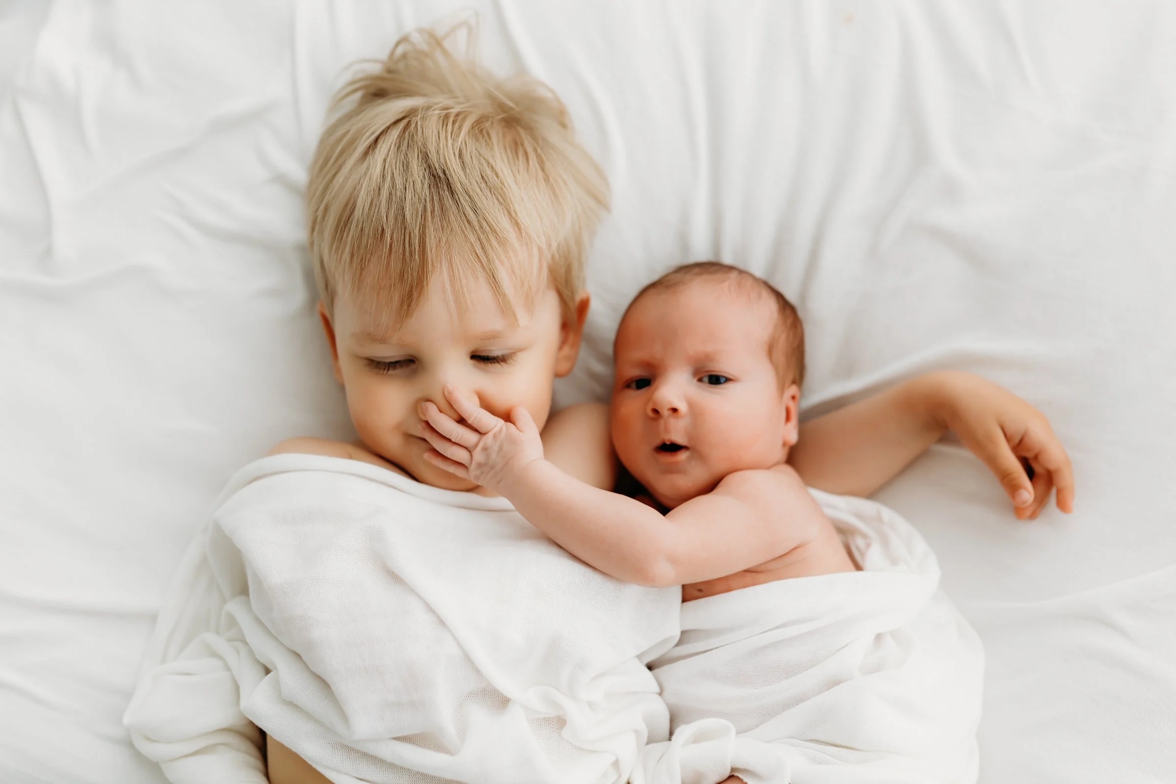 A young boy with blond hair and his infant sibling lying together on a bed, both wrapped in white blankets. The boy has his eyes closed, while the baby appears to be looking at the boy with a surprised expression, touching the boy's face.