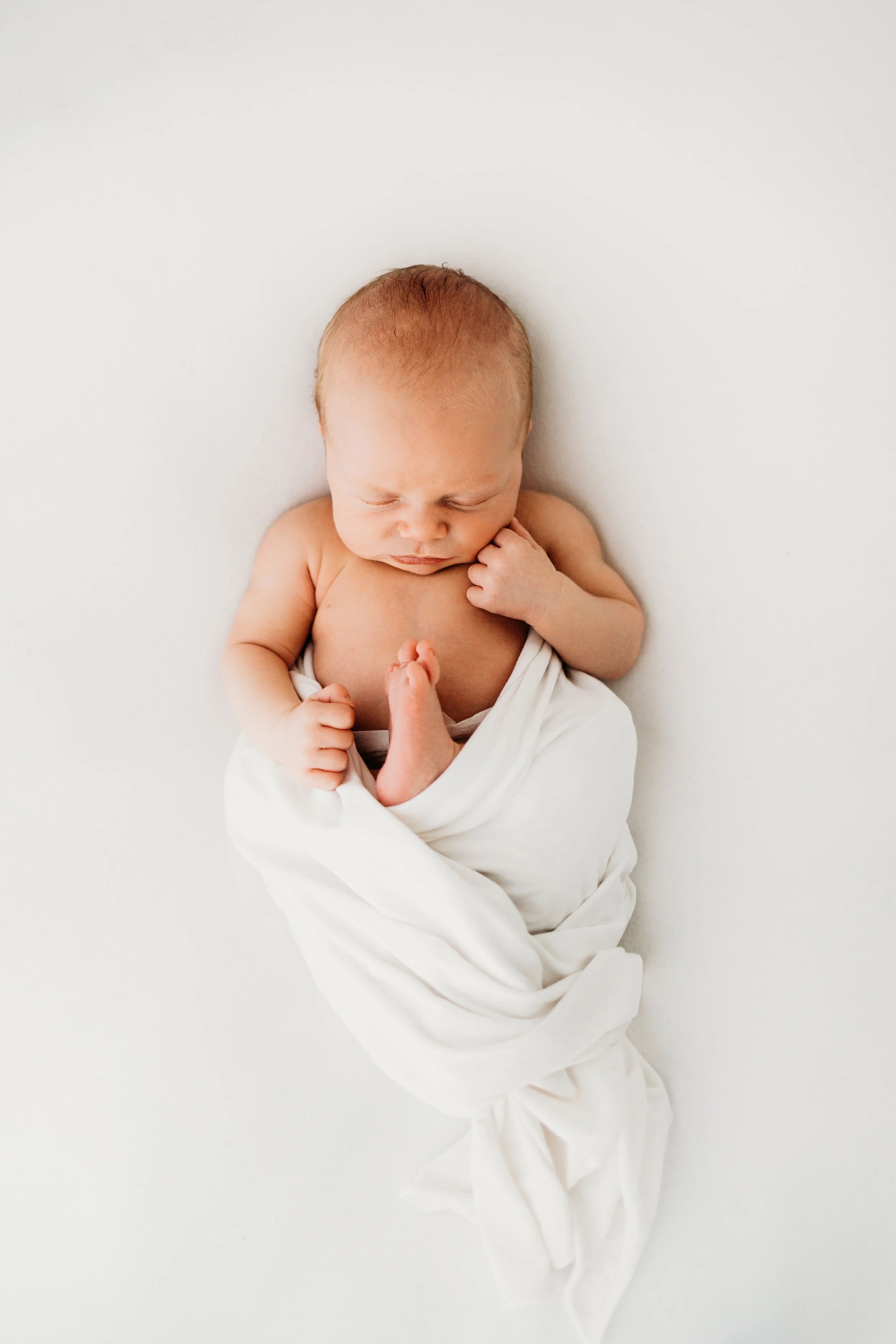 Newborn baby with fair skin and light brown hair, wrapped in a white cloth, lying on a white surface, with one hand near the face and the other holding the foot.