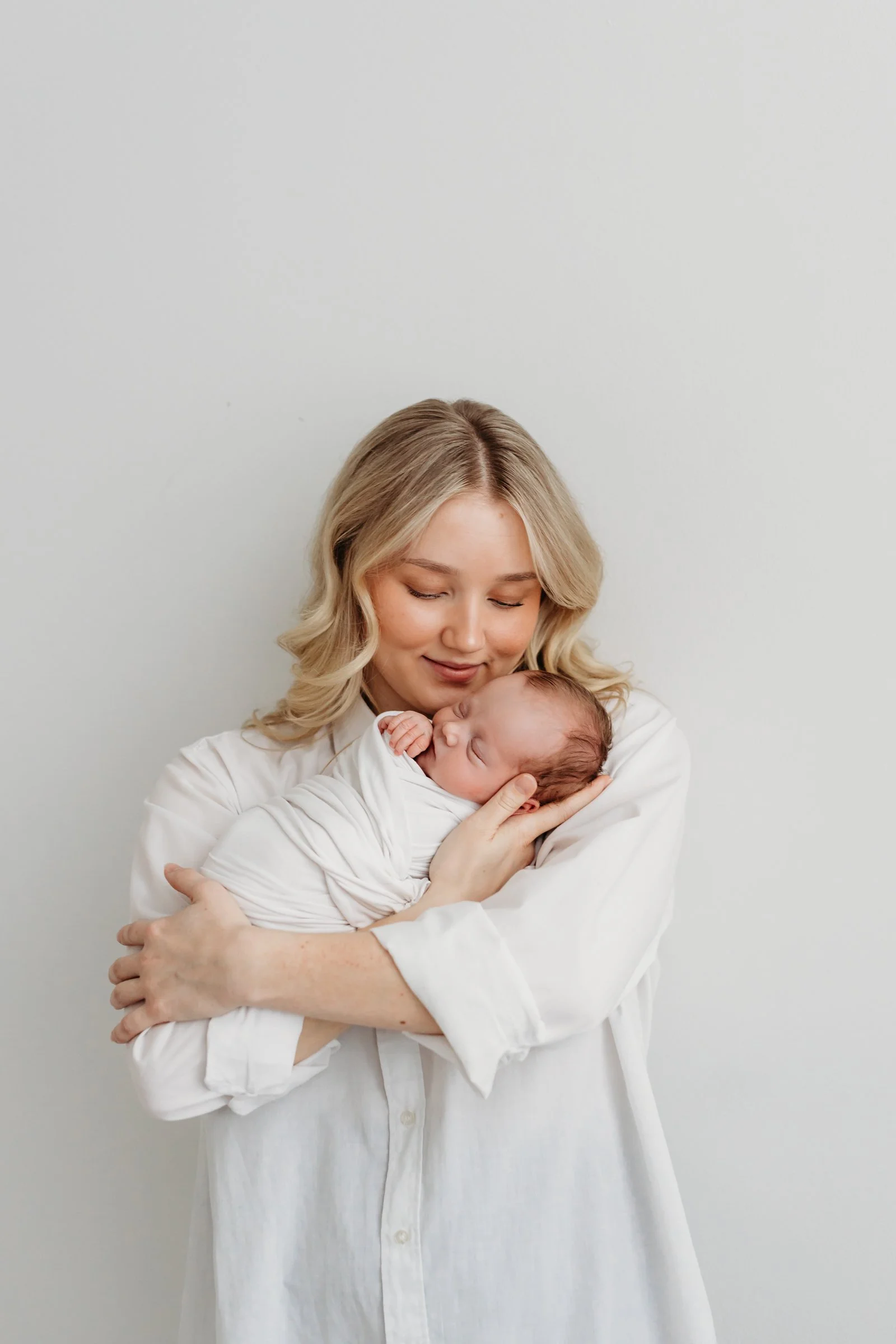 A woman with blonde hair holding a newborn baby, both dressed in white, against a plain white wall.