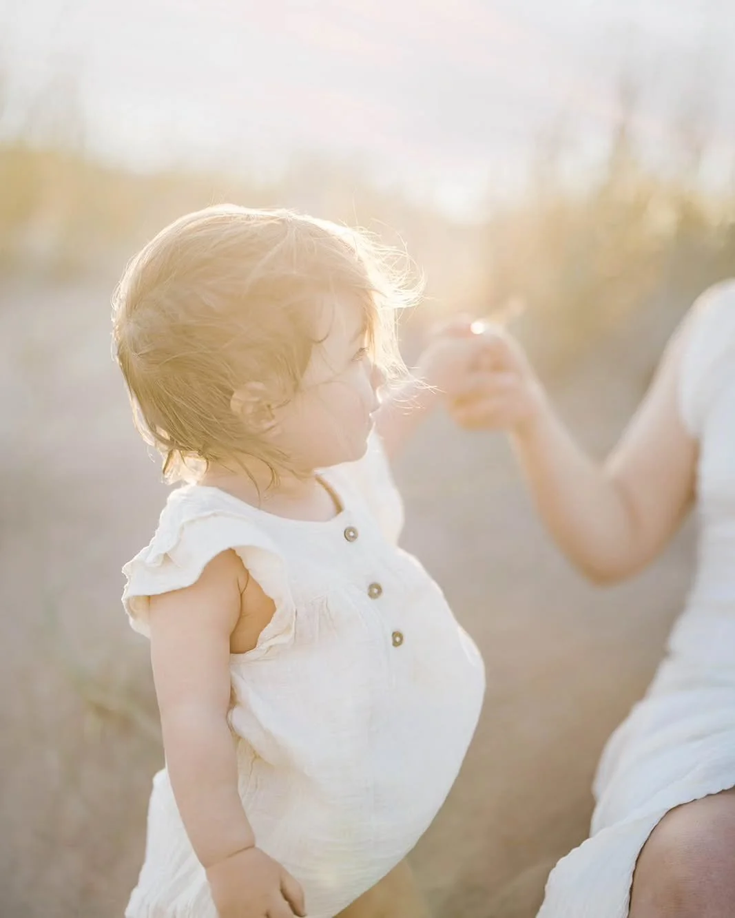A young girl with short, curly hair wearing a white dress standing outdoors in sunlight.
