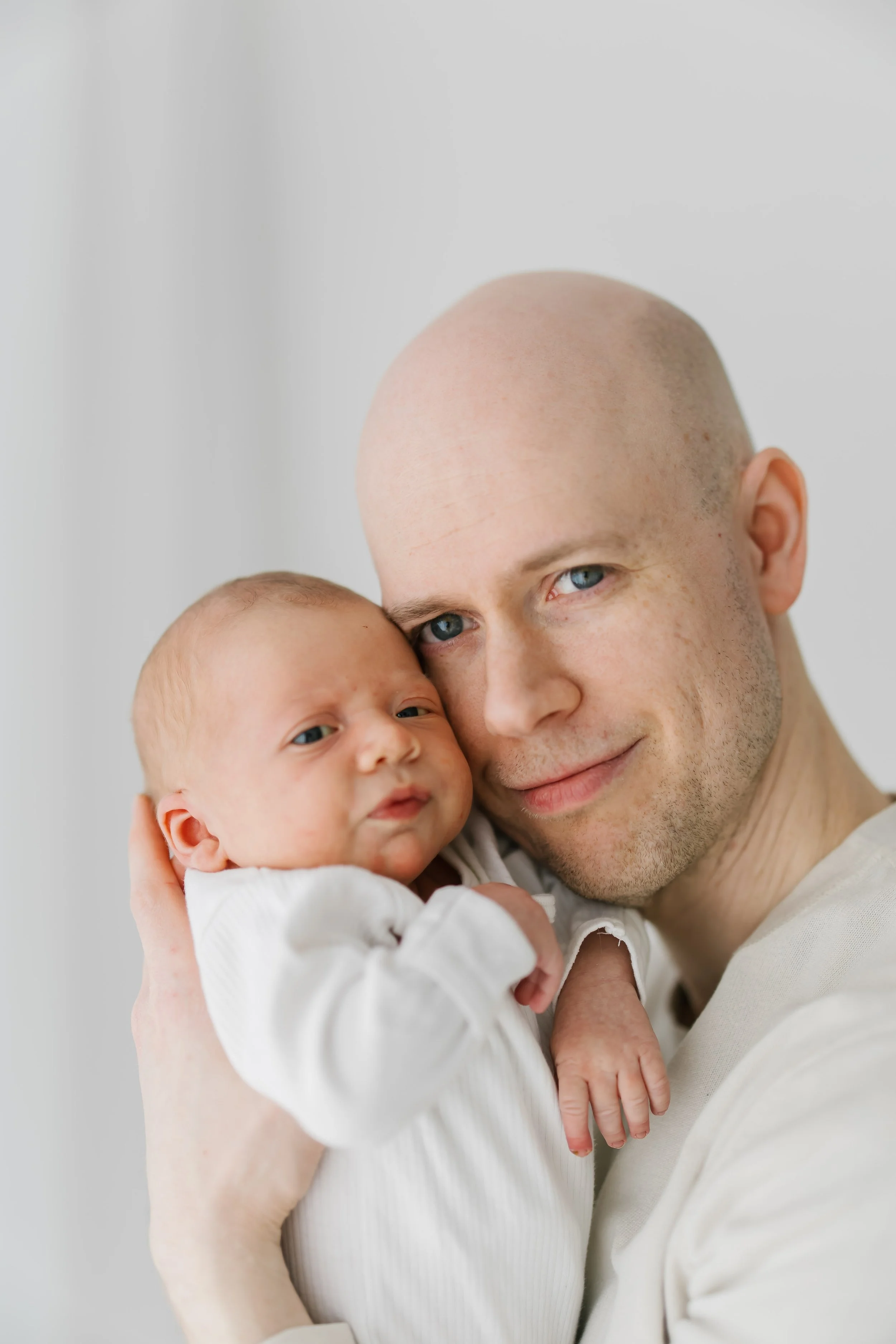 A man holding a newborn baby close to his face, both looking at the camera. The man has a shaved head and blue eyes, and the baby is wearing a white outfit with a serious expression.