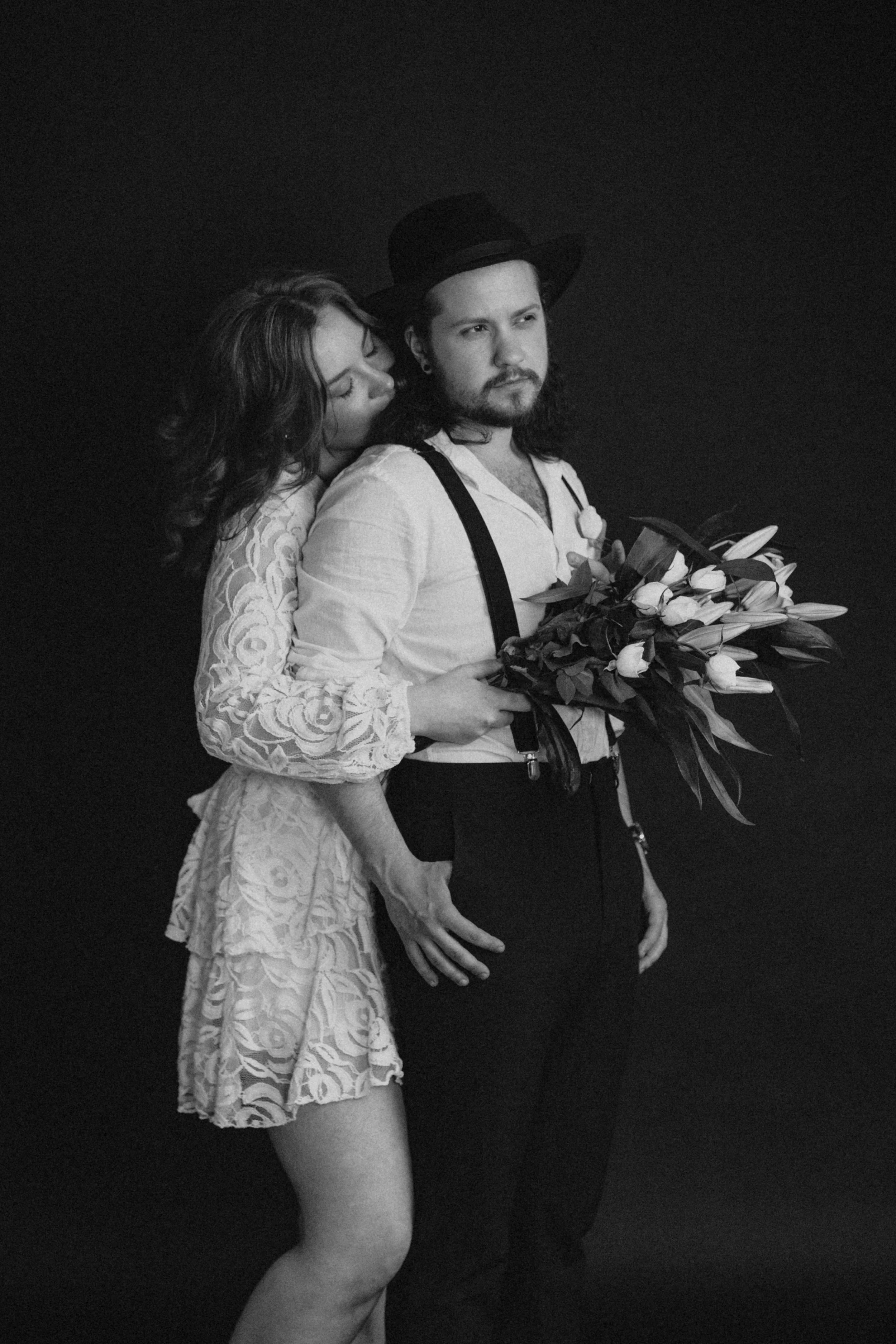 A black-and-white photo of a woman embracing a man holding a bouquet of flowers, with the woman leaning her head on the man's shoulder. The man has a beard, long hair, and is wearing a hat, suspenders, and an open shirt. The woman has curly hair and is wearing a lace dress.