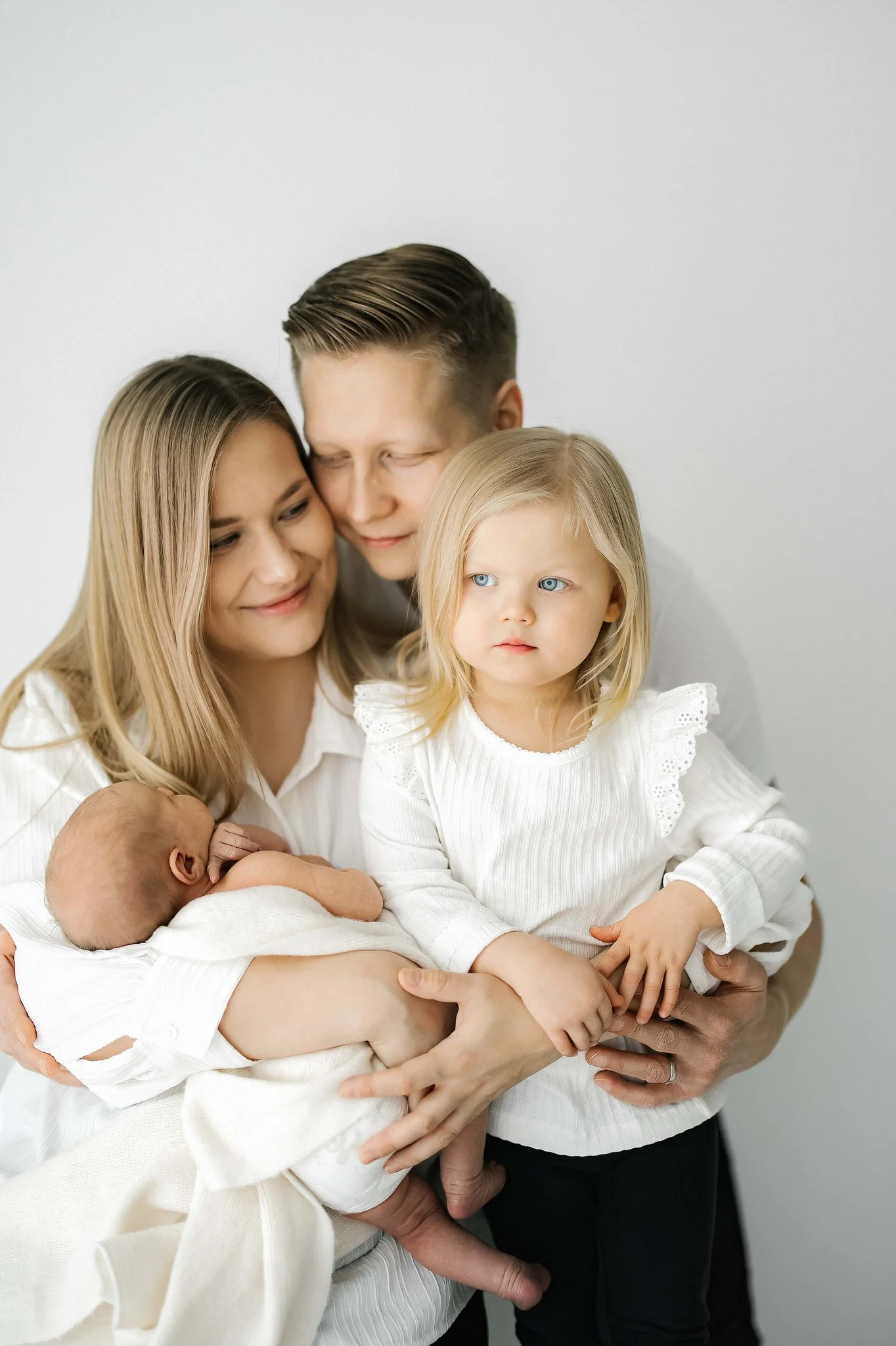 A family of five in white clothing, including a baby, a young girl, and two adults, standing close together against a plain white background.