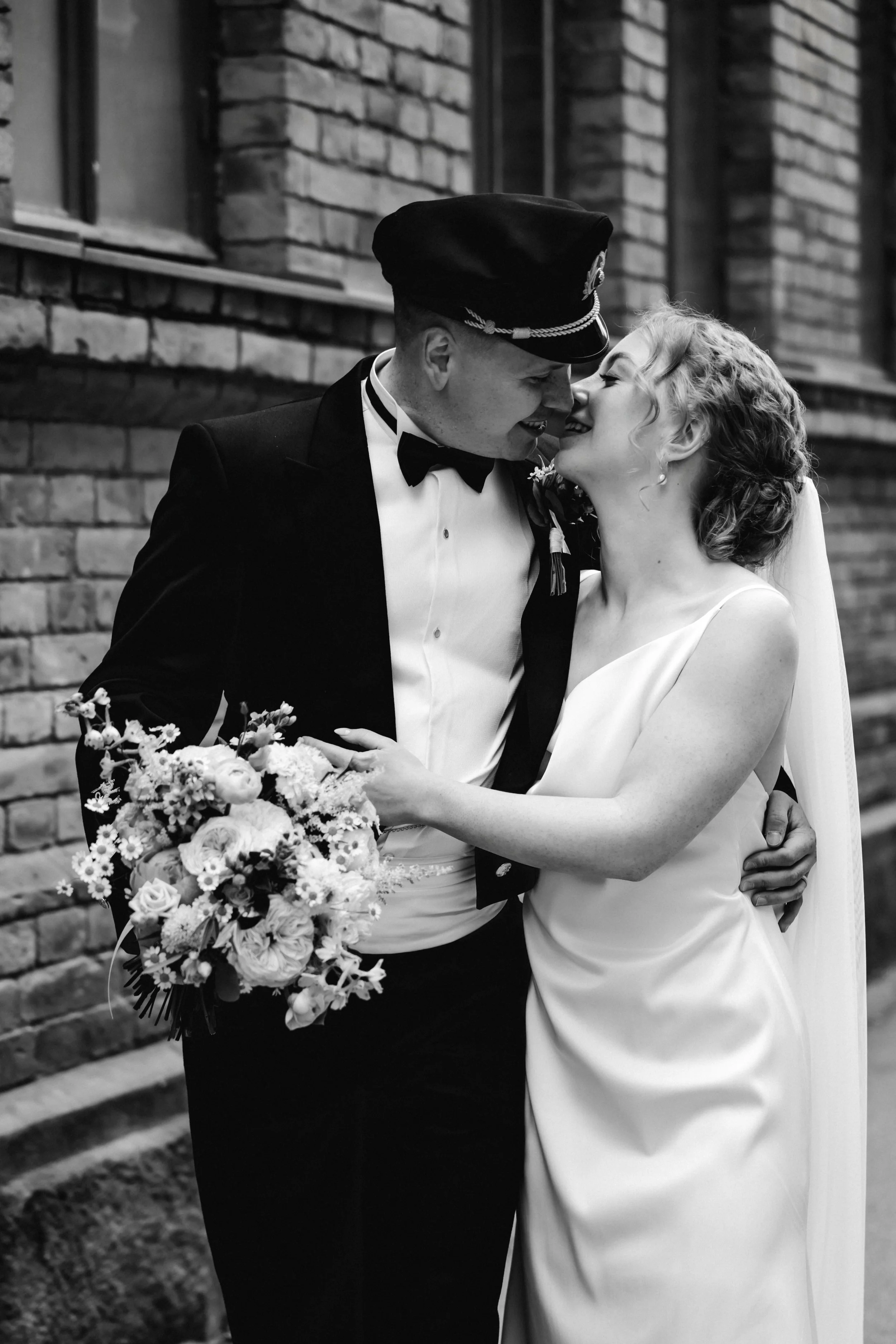 A black and white photo of a bride and groom sharing a kiss outdoors, with the bride holding a bouquet of flowers and wearing a wedding dress, while the groom is dressed in a tuxedo with a hat, standing against a brick wall.