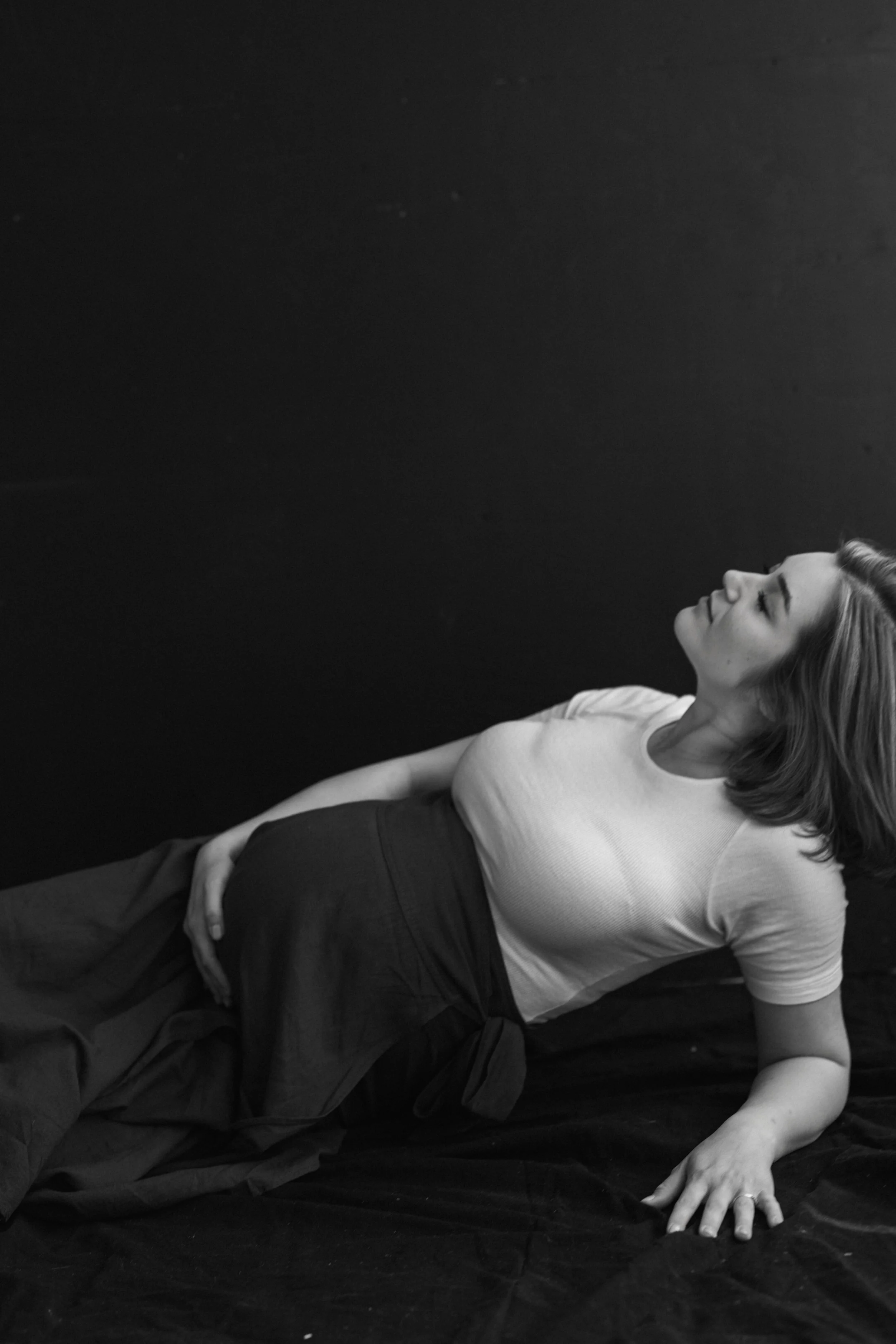 A woman lying on a dark surface with her eyes closed, wearing a white t-shirt and dark pants, with one hand resting on her side and the other on the surface.