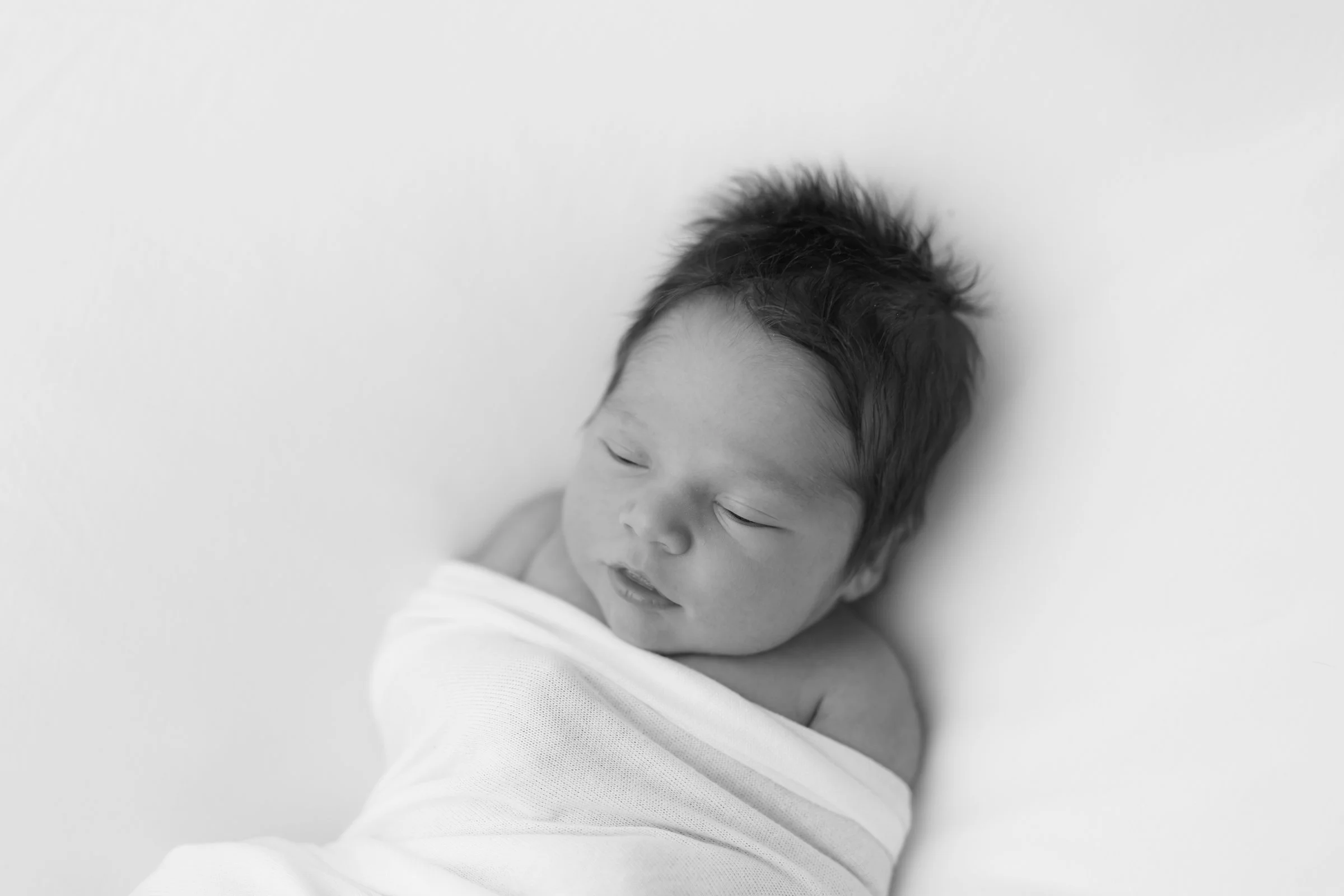 Black-and-white photo of a newborn baby wrapped in a white blanket, sleeping on a plain white surface.