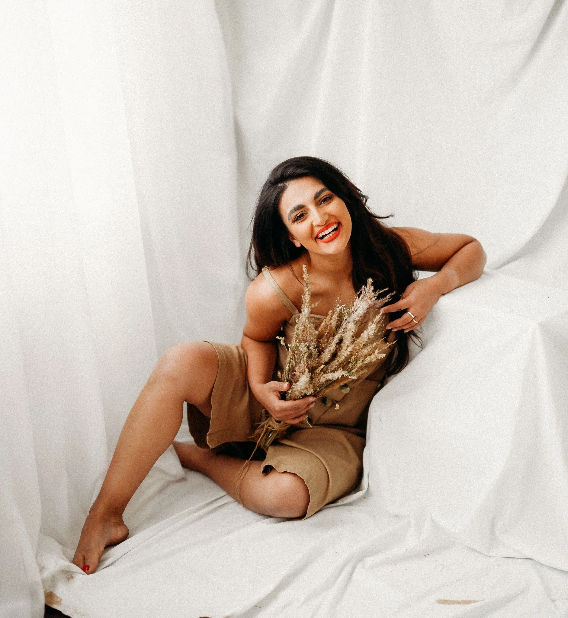 Smiling woman in beige clothing sitting on white sheets, holding a bouquet of dried flowers.