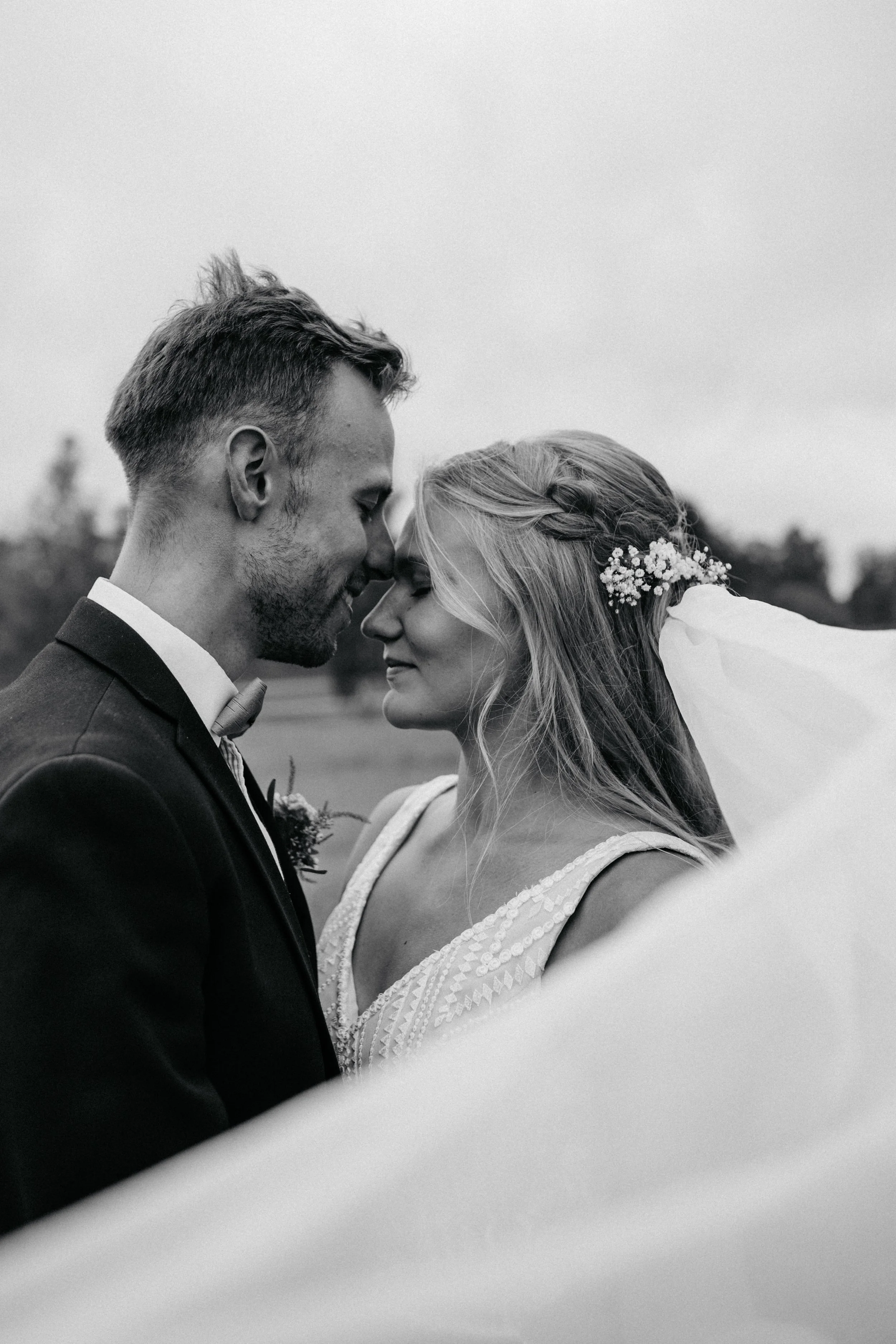 Black and white photograph of a bride and groom with foreheads touching, eyes closed, smiling softly outdoors.