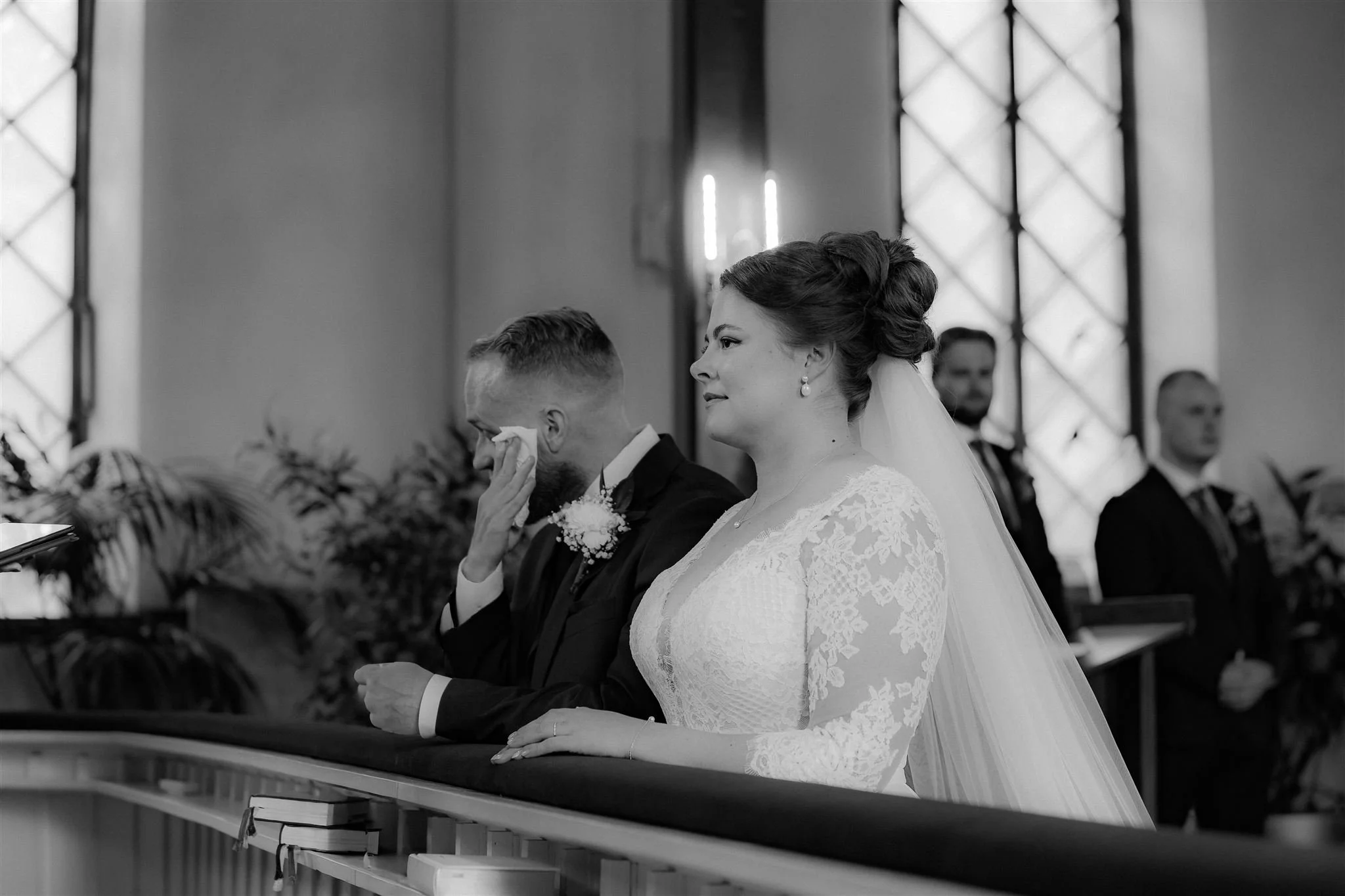 A bride and groom stand at an altar during their wedding ceremony, with the groom wiping away tears and the bride looking solemn, in a church setting.