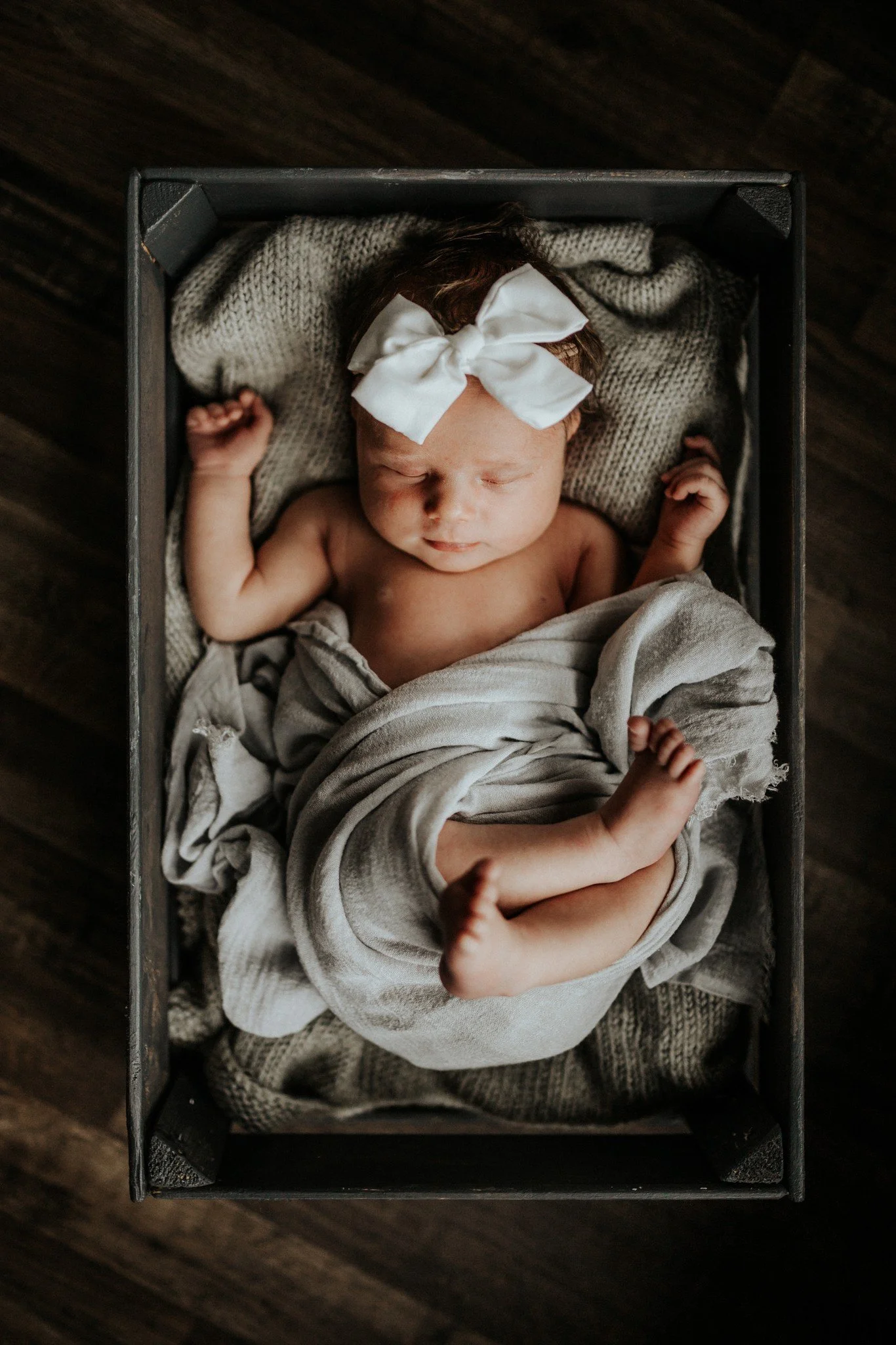 A baby girl sleeping in a cardboard box, wearing a large white bow headband and wrapped in a beige blanket, lying on a knitted blanket with hardwood flooring visible around.