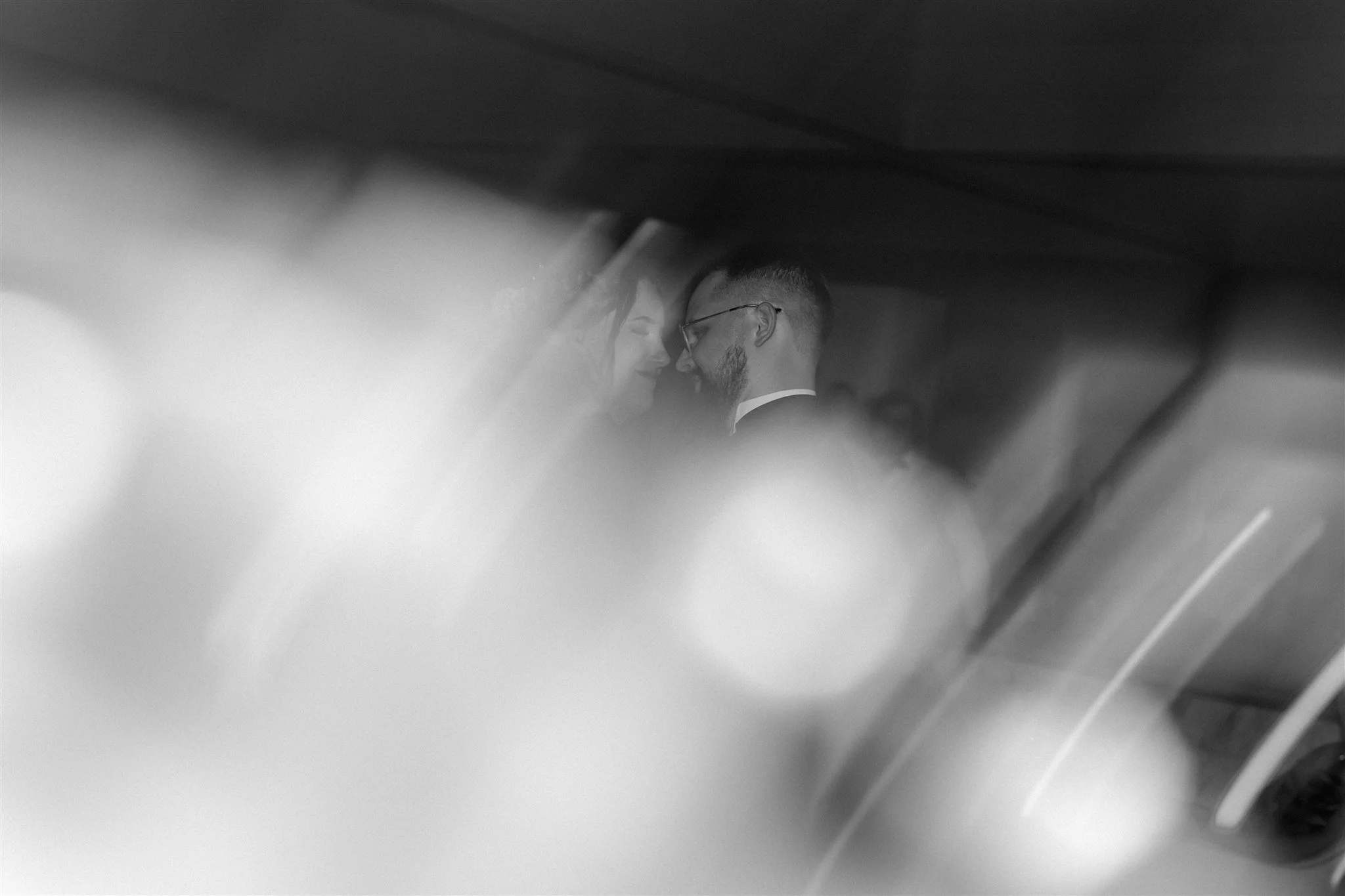 Black and white photo of a couple touching foreheads, with soft focus and blurred foreground elements.