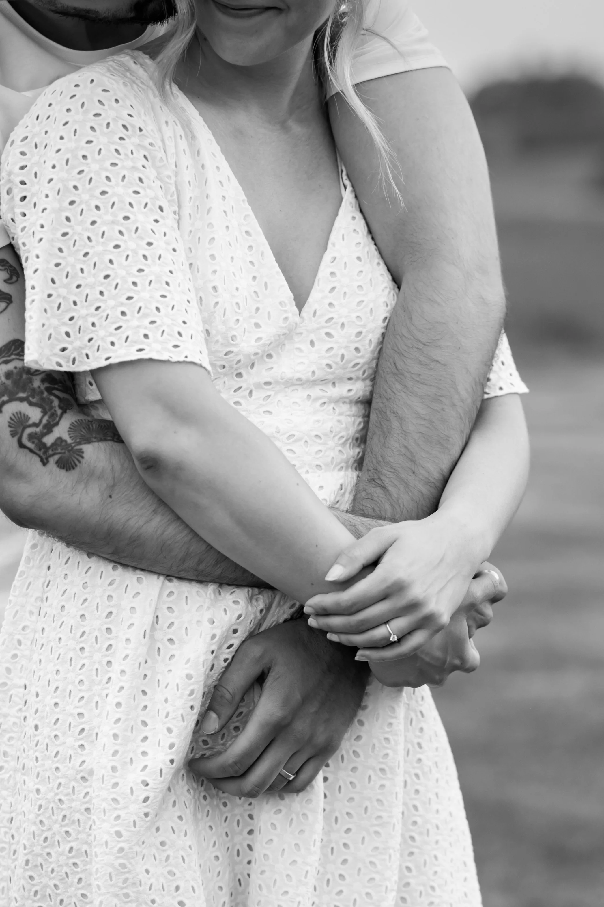 A close-up black and white photo of a couple embracing, focusing on their hands and arms. The woman is wearing a light-colored dress with a eyelet pattern, and the man has a tattoo on his arm. The woman is wearing a ring on her finger.