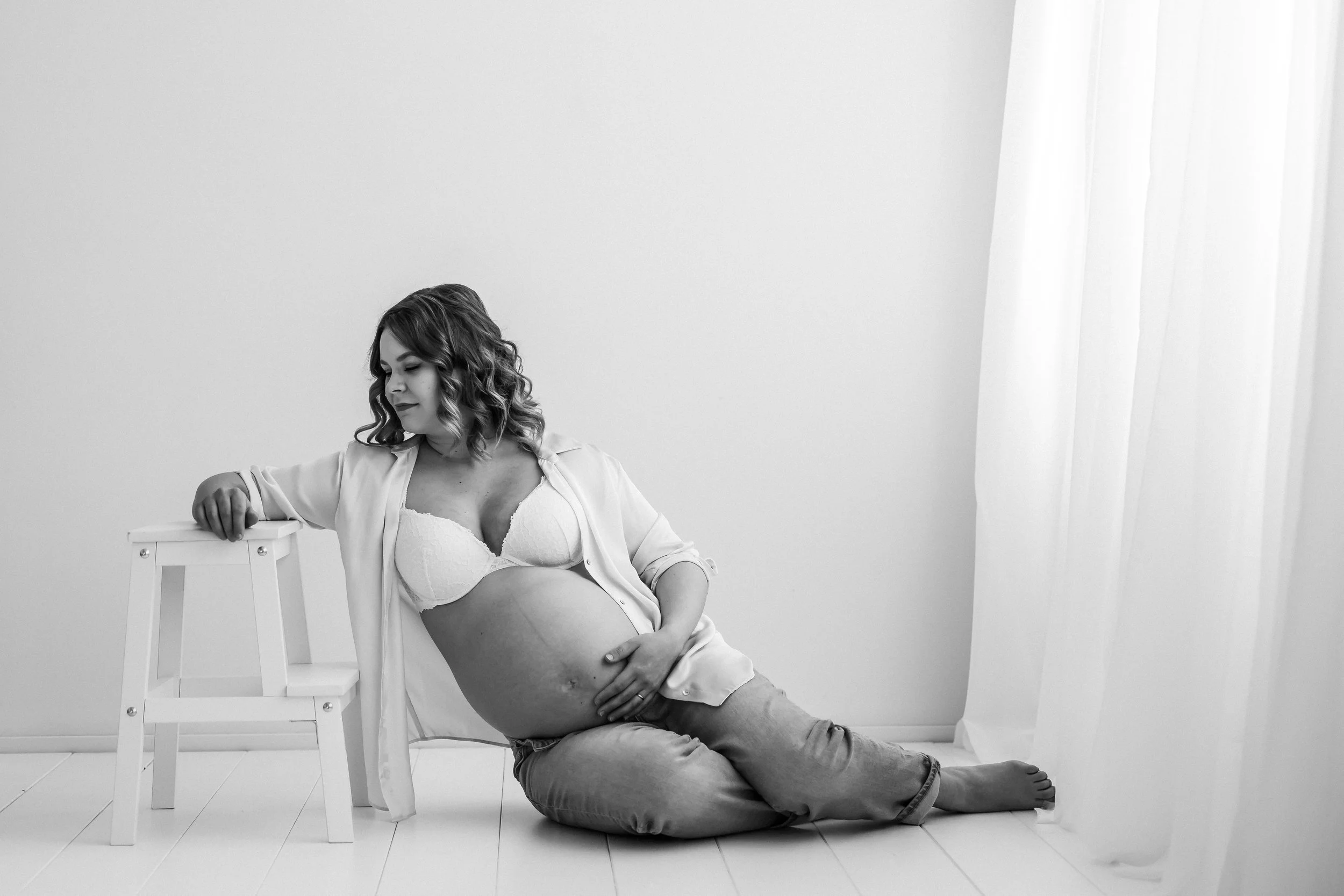 Black and white photograph of a pregnant woman sitting on the floor, leaning against a small white stool, wearing a bra and open shirt, with her hand resting on her belly and her eyes closed.