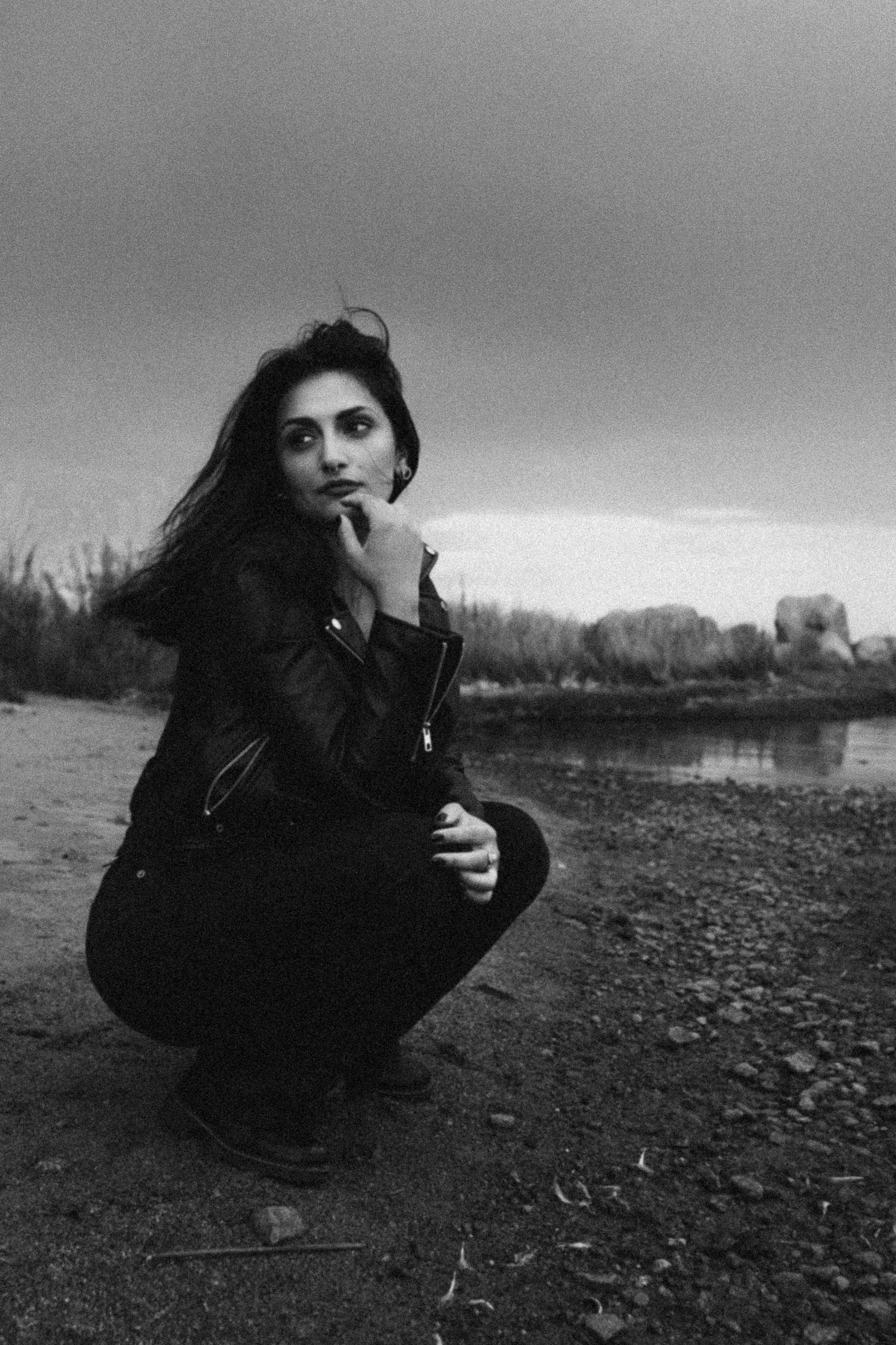 A woman with dark hair wearing a leather jacket, squatting on a sandy beach with water and trees in the background, captured in black and white.