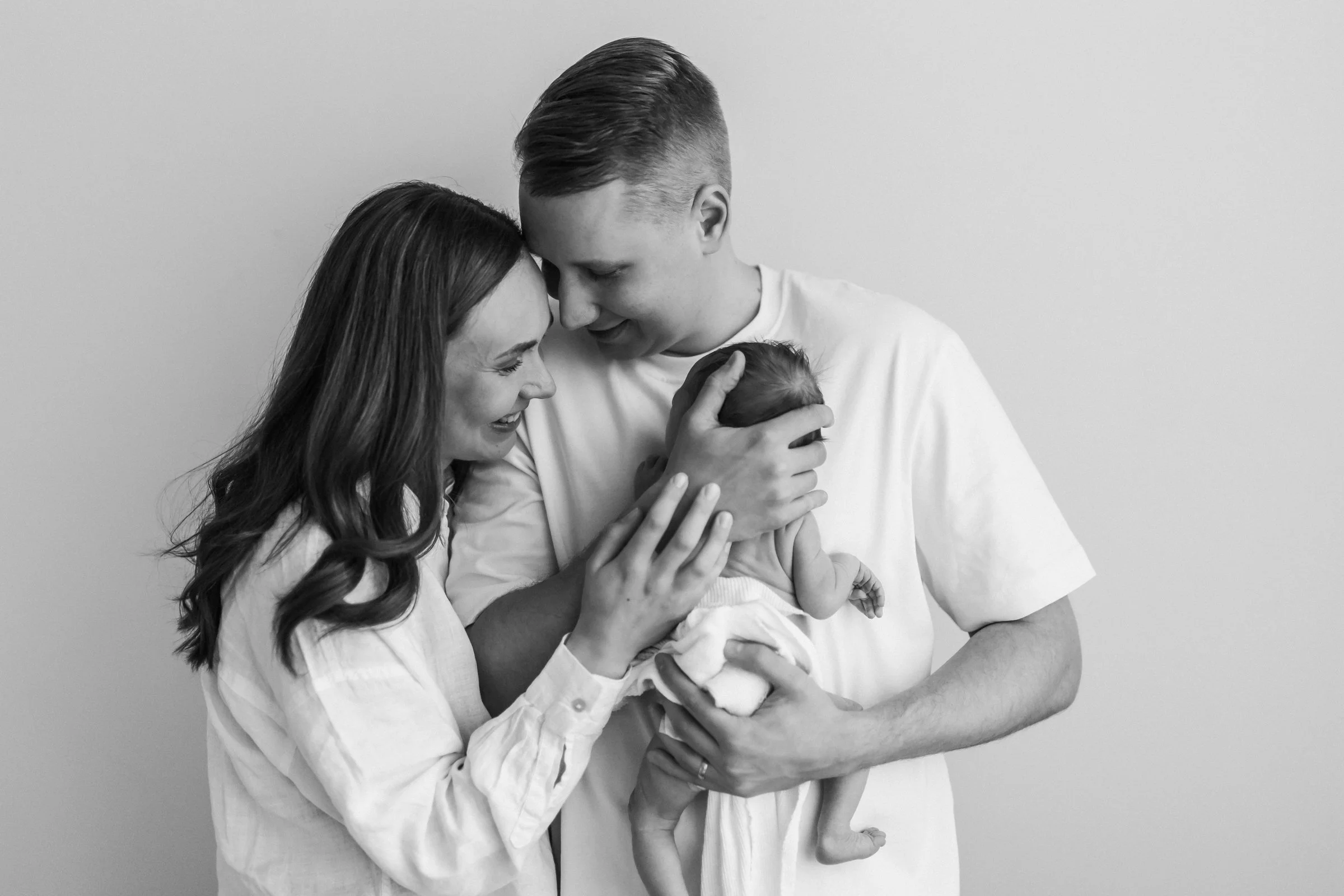 A black and white photo of a family holding a newborn baby. The woman and the man are smiling and touching their foreheads together while looking at the baby, who is cradled in the man's arms.