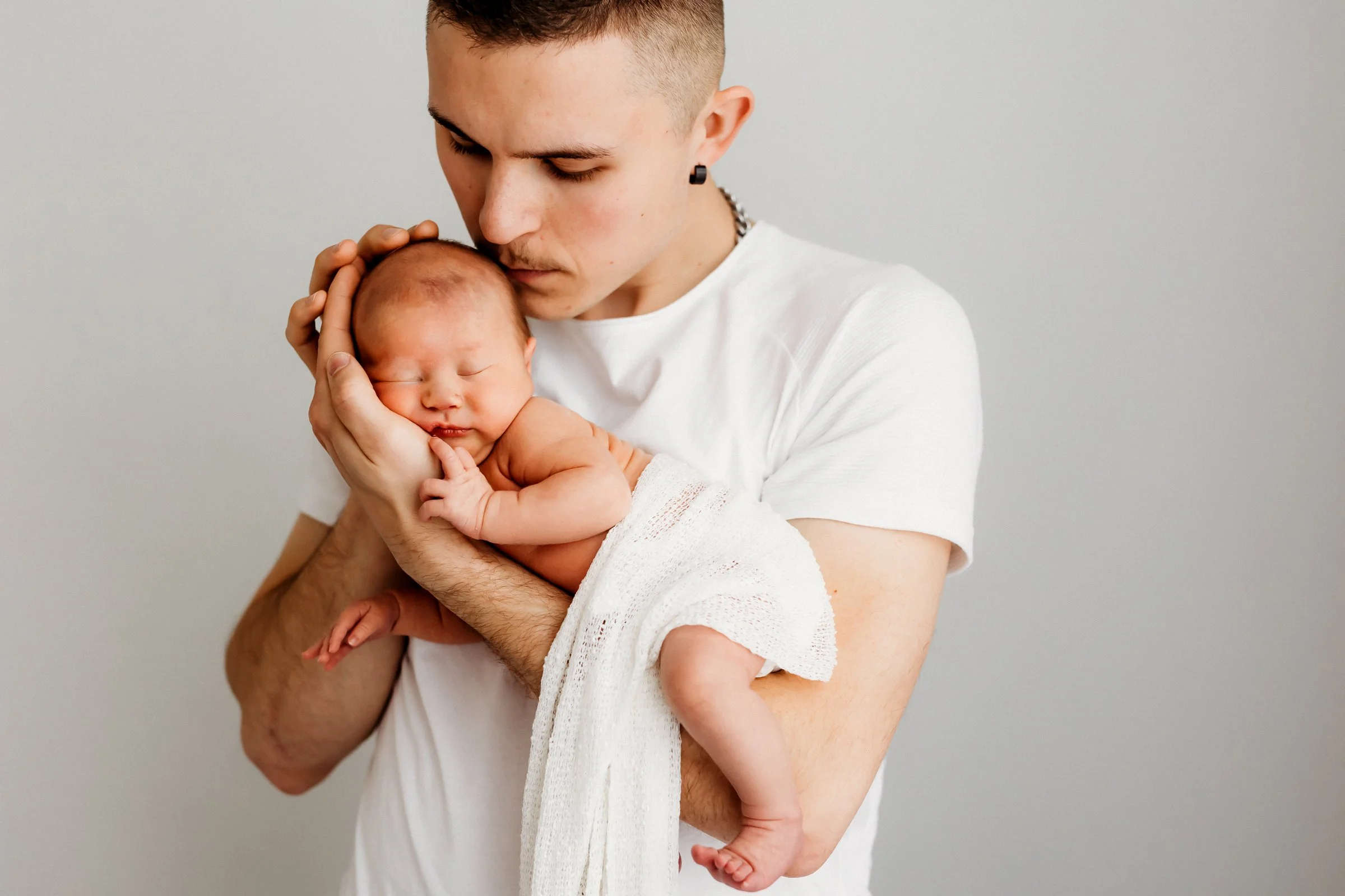 A young man holding a sleeping newborn baby close to his face against a plain, light gray background.
