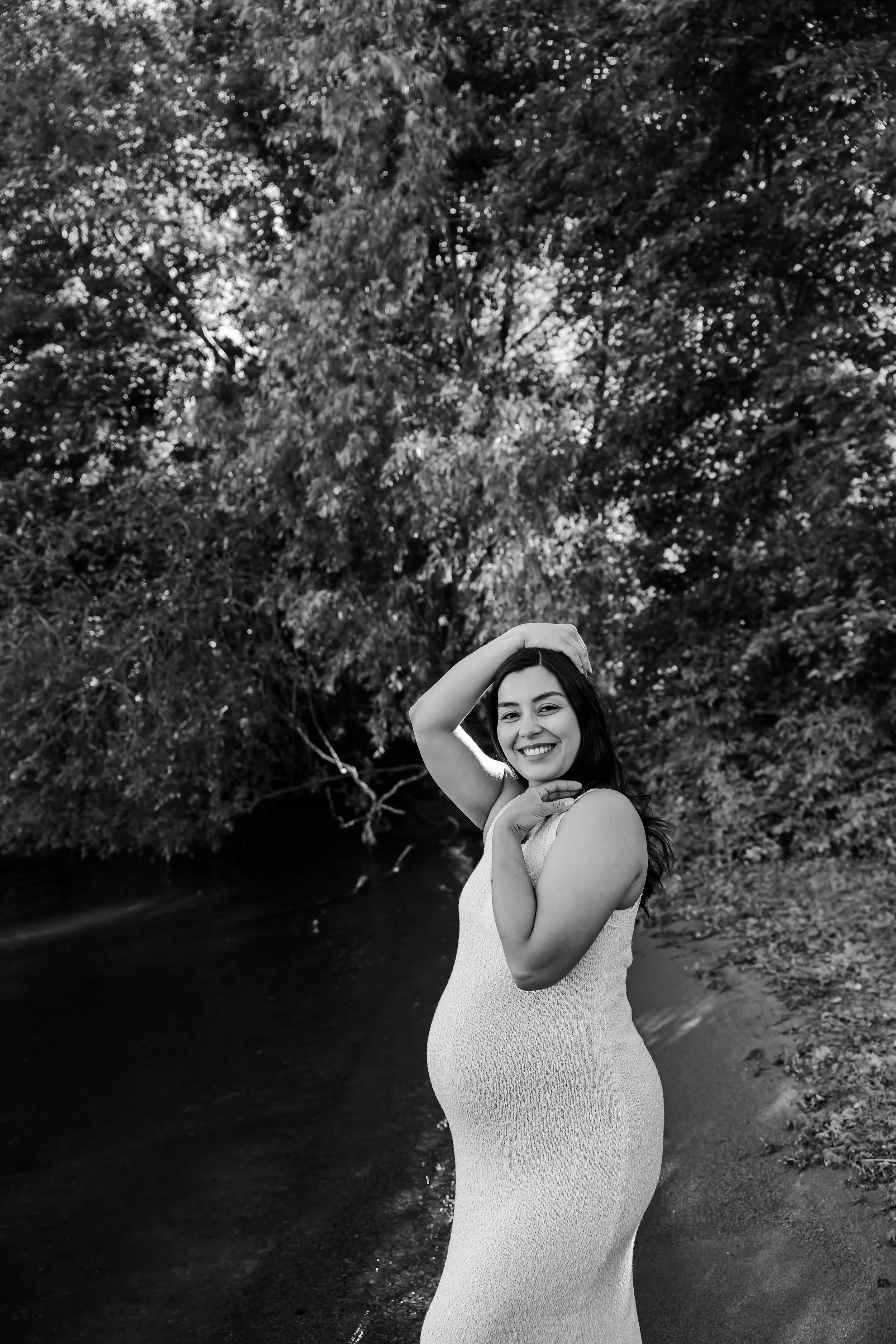 A woman in a sleeveless dress smiling and posing on a riverbank surrounded by trees.