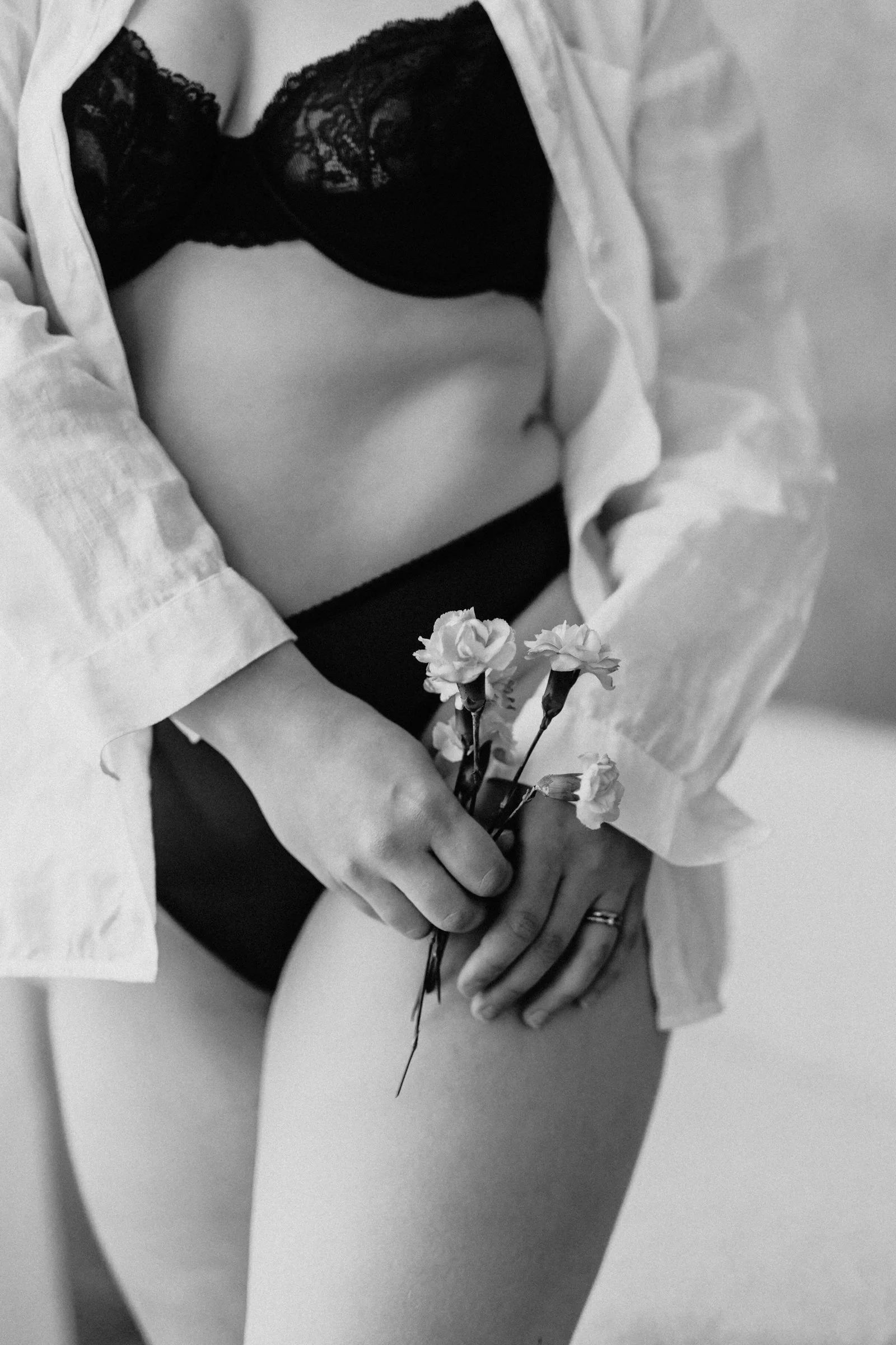 A person wearing black lingerie and an unbuttoned shirt, holding small flowers with one hand over their thigh.