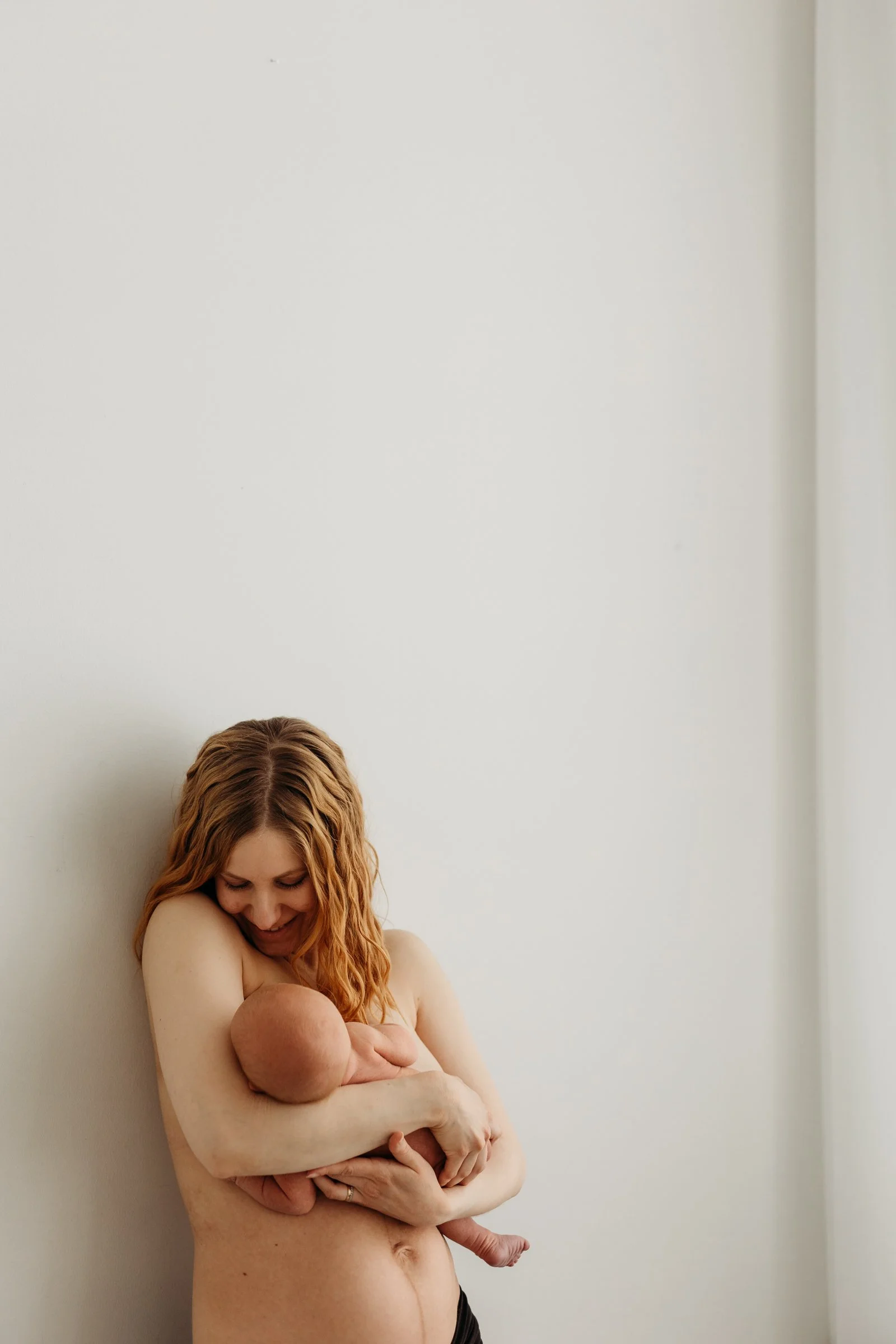 A woman with red hair breastfeeding a baby against a plain white wall.