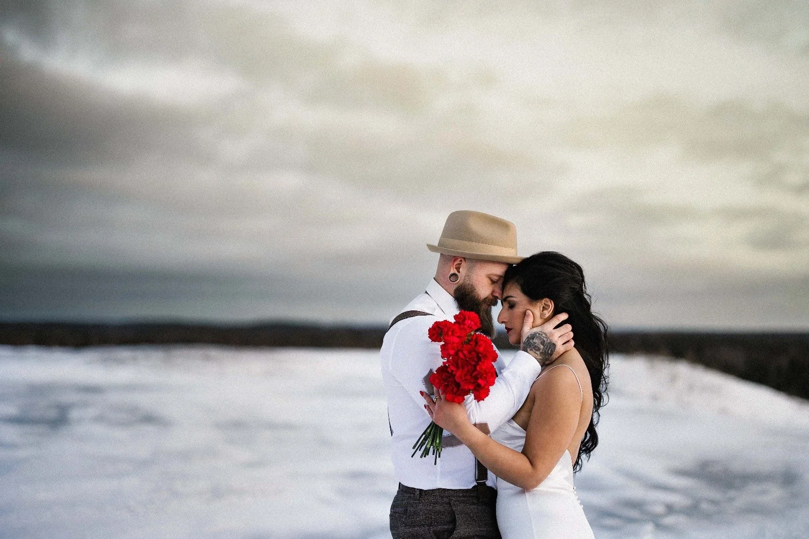 A couple in wedding attire embracing outdoors on a snowy landscape under a cloudy sky, the man holding a bouquet of red flowers.