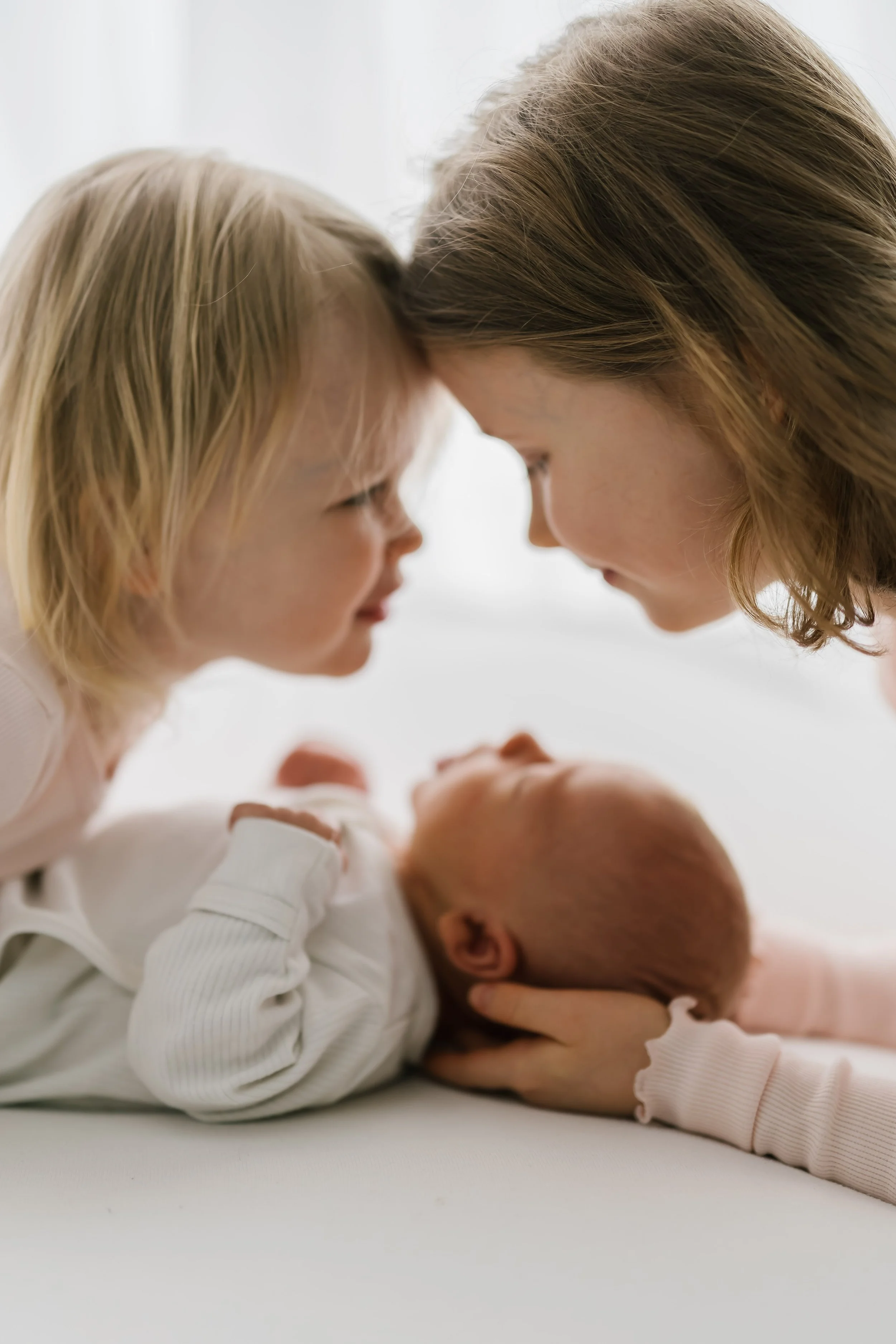 A woman and a young girl touching foreheads above a newborn baby lying on a white surface.