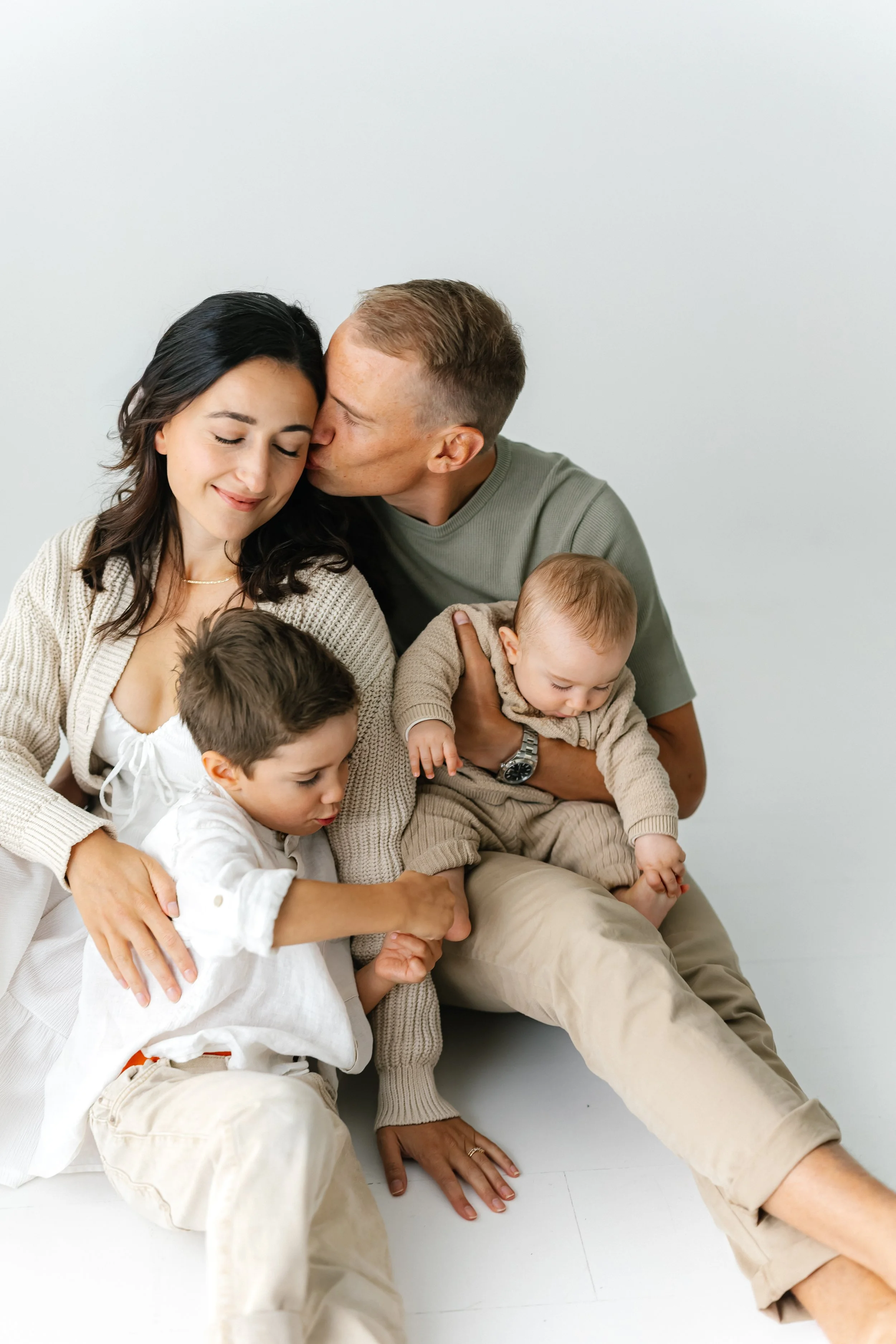 A family of four, including a woman, a man, and two young boys, sitting close together on the floor in a minimalistic white room. The man is giving a kiss on the woman's cheek while the woman smiles with eyes closed, and one boy is grabbing the woman's knee while the other is being held by the man.