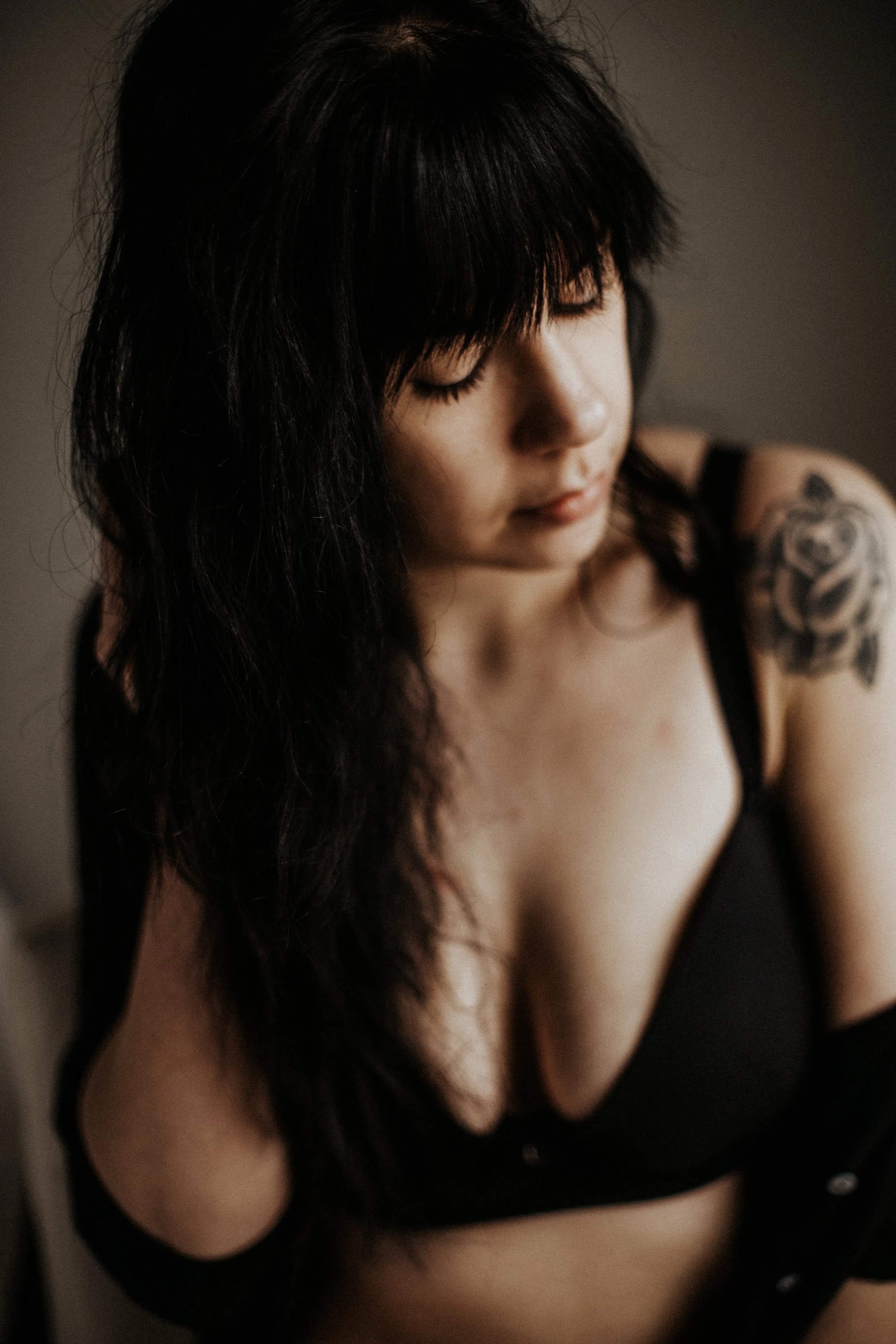 A woman with black hair and a tattoo on her shoulder, wearing a black top, looking down with closed eyes.