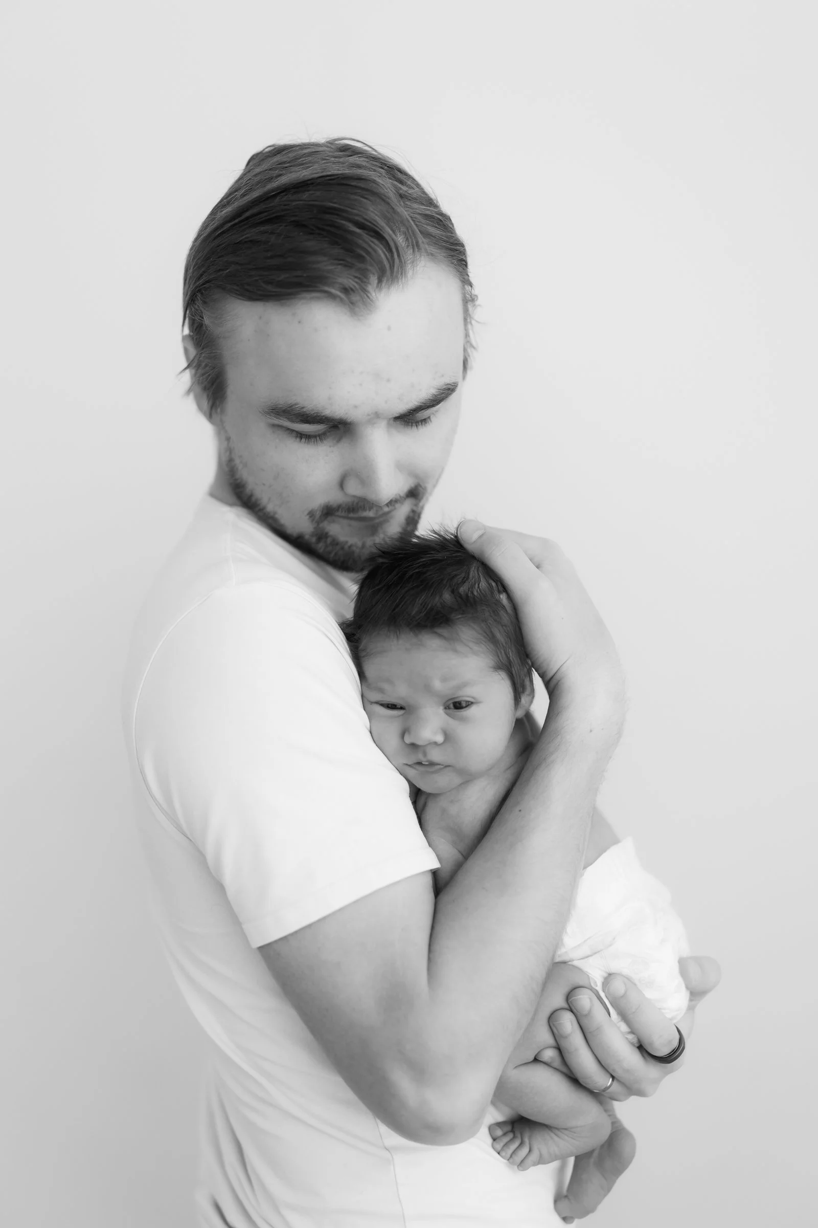A man with a beard holding a newborn baby close to his chest, both facing downward, against a plain white background.
